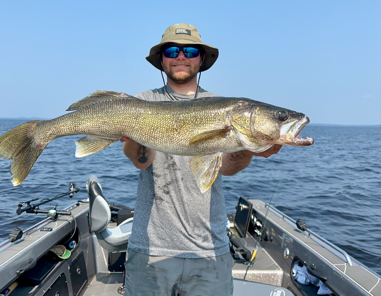 A man standing on a boat holding a large fish with both hands, with water and a clear sky in the background.