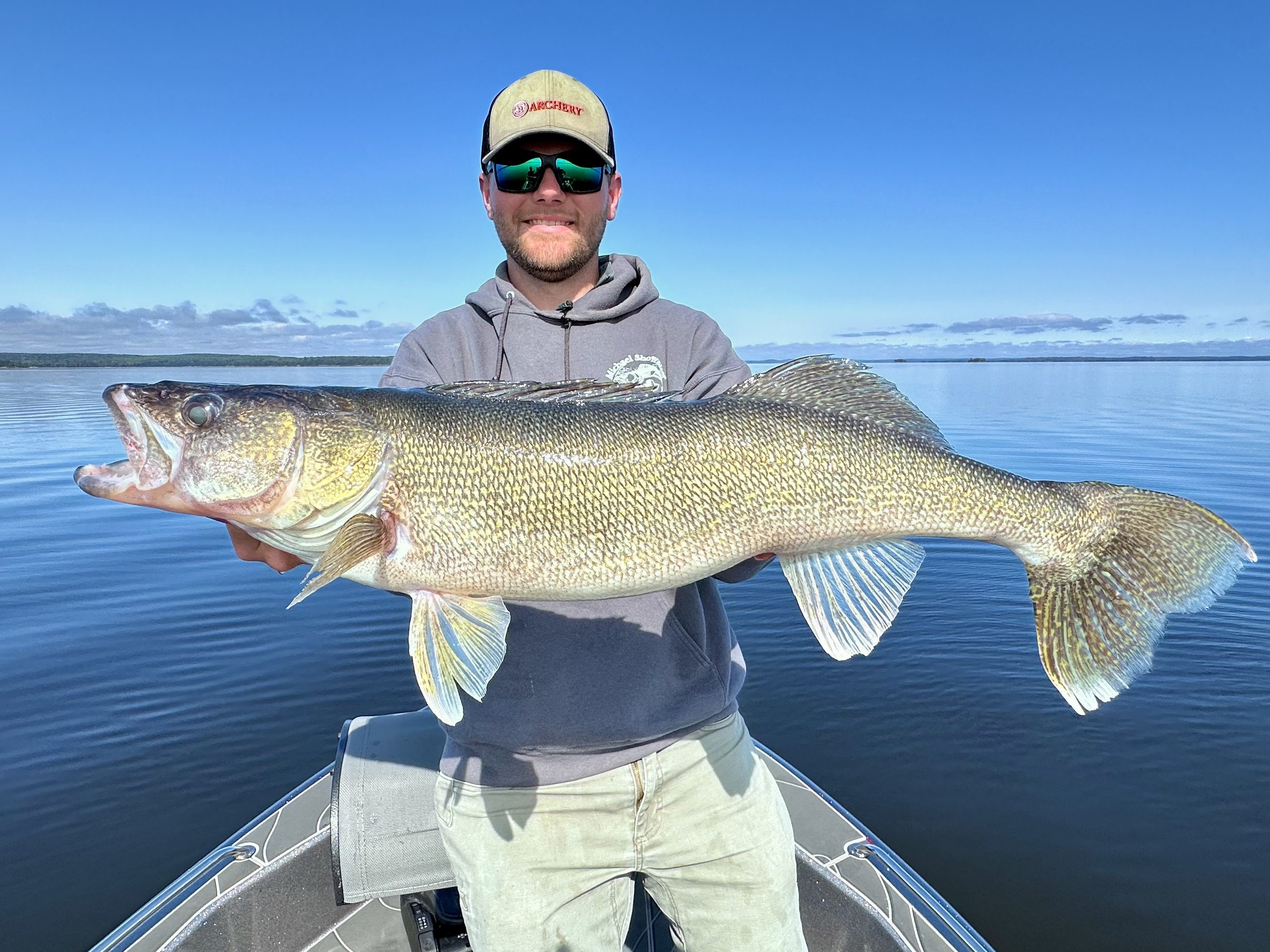 Man in hoodie and sunglasses holding large fish on boat with calm lake and blue sky background.
