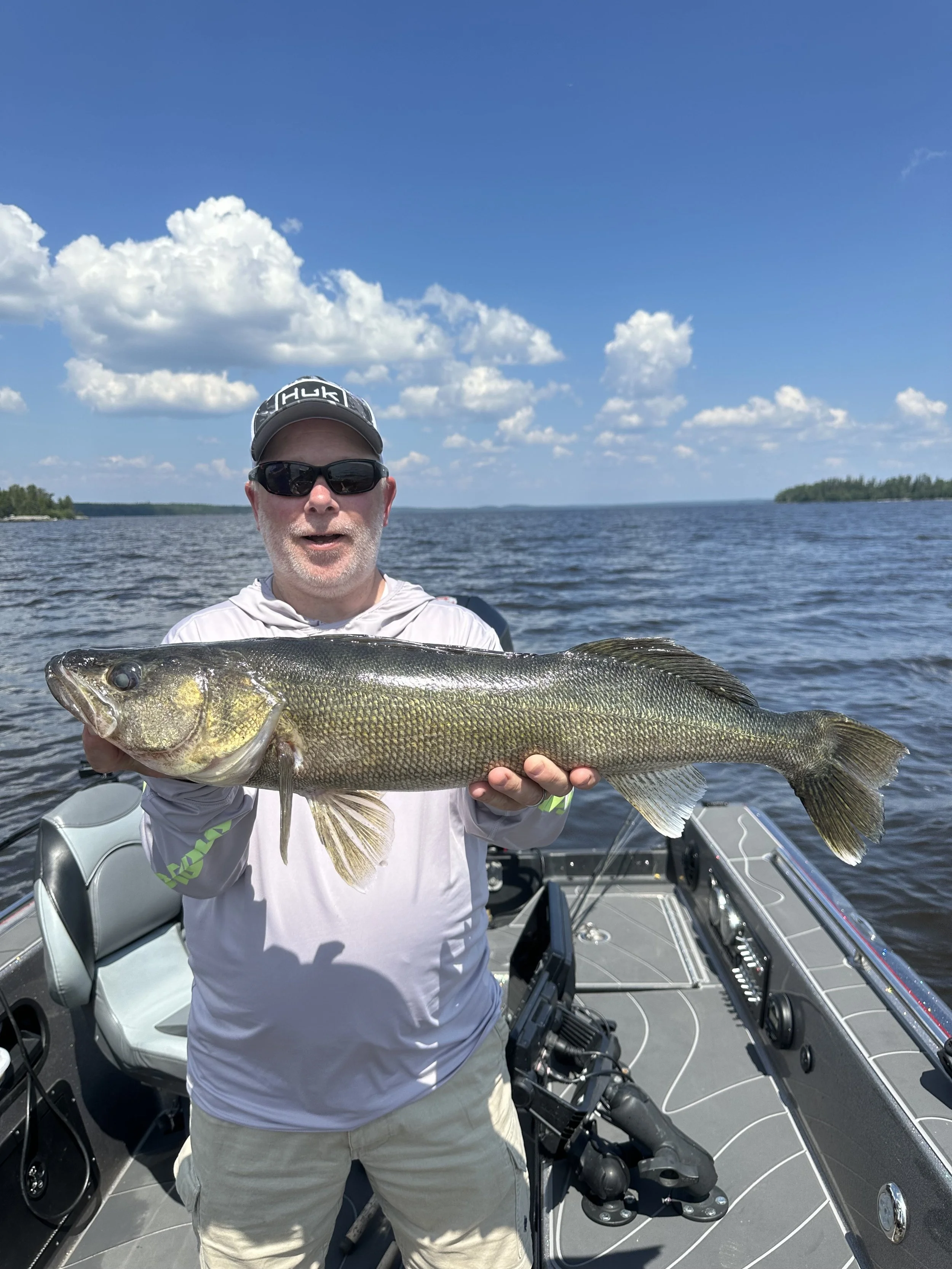 Man wearing a gray long-sleeve shirt, sunglasses, cap, holding a large fish on a boat in a lake, with a background of water and partly cloudy sky.