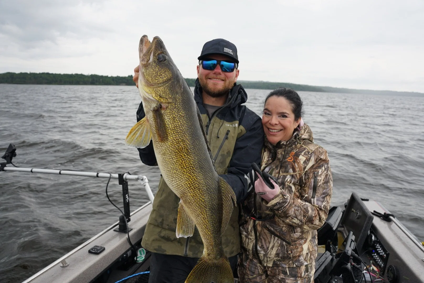 A man and woman on a boat holding a large fish, with the woman smiling and making a peace sign, in a body of water with a cloudy sky in the background.