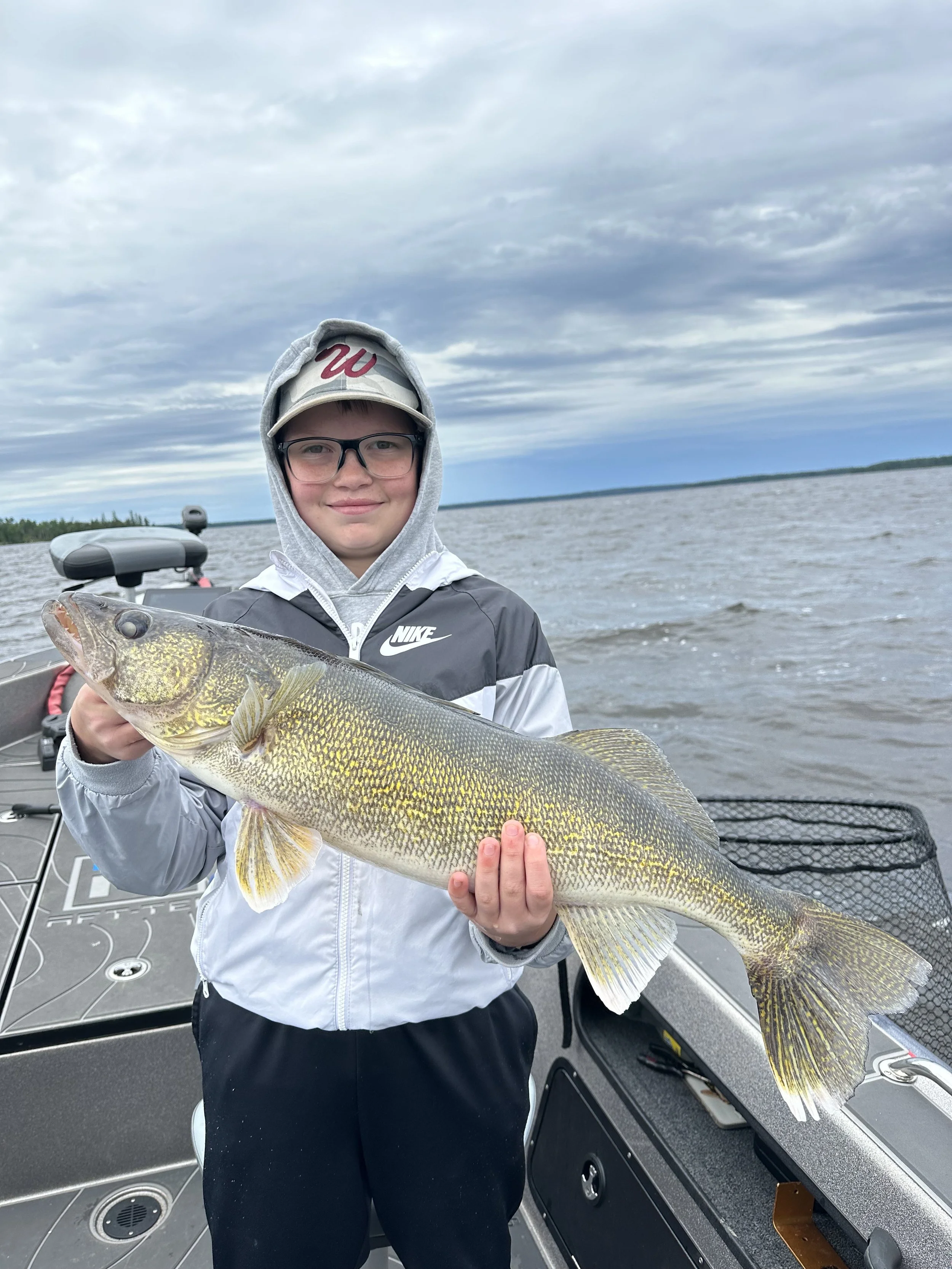 A young boy wearing glasses, a gray hoodie under a Nike jacket, and a baseball cap, holding a large fish on a boat in a lake with cloudy skies in the background.