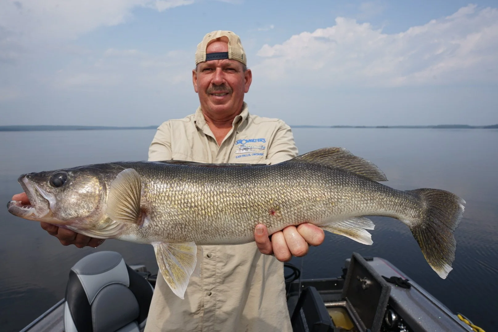 Man holding a large fish on a boat, with a body of water and a partly cloudy sky in the background.