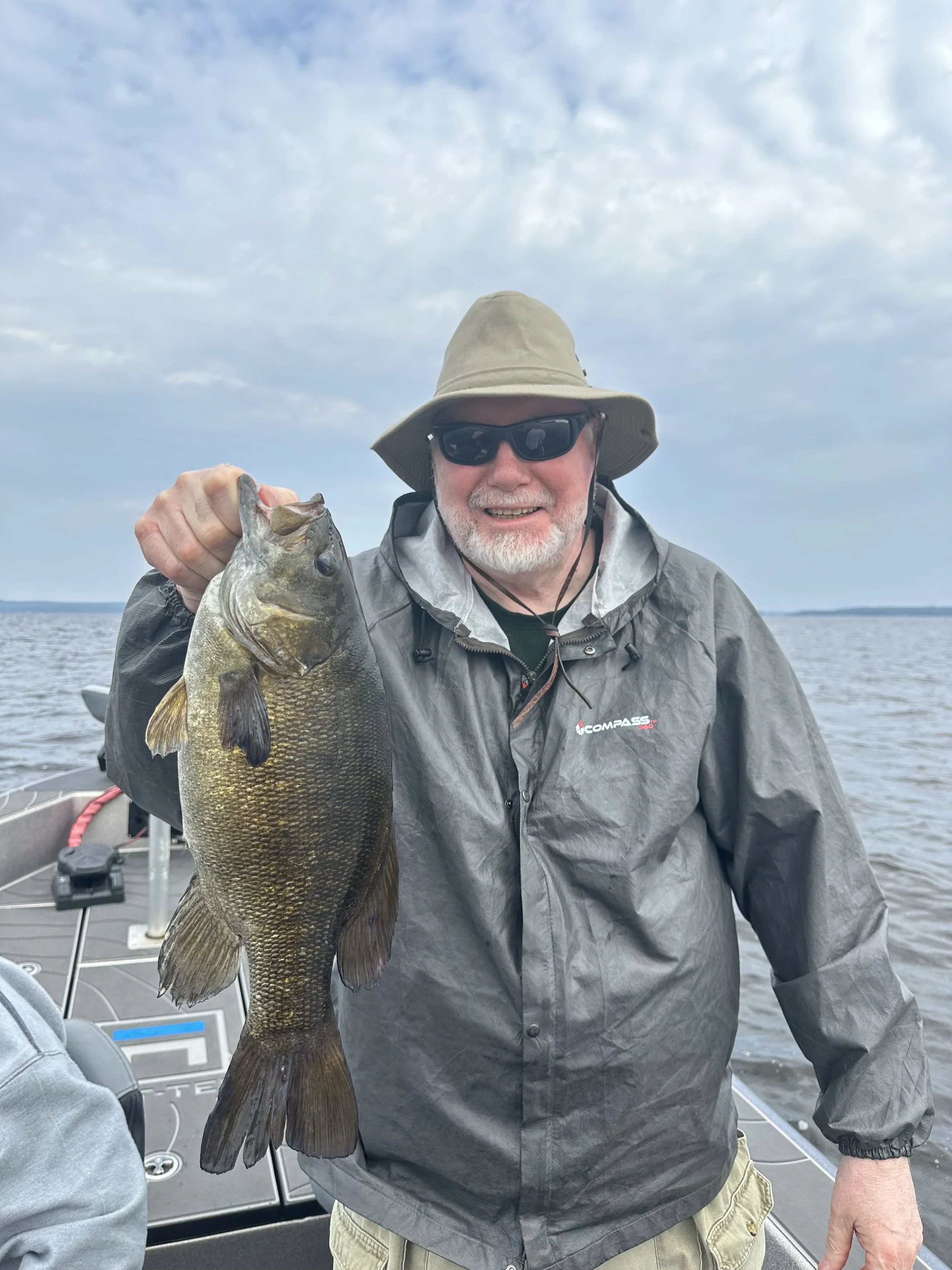 A man wearing a wide-brimmed hat, sunglasses, and a gray jacket, smiling while holding a large fish on a boat in a lake with a cloudy sky.