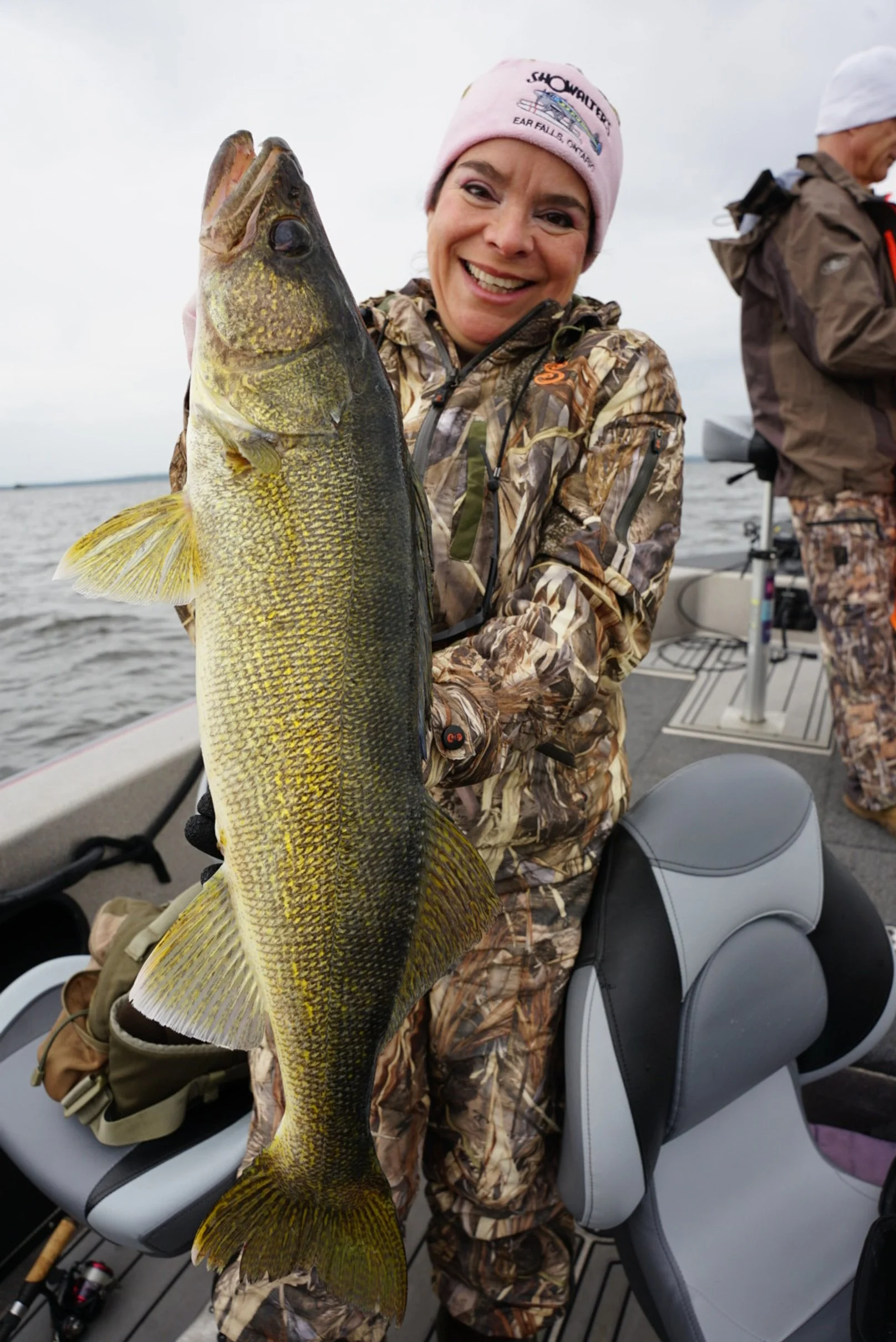 A woman in camouflage clothes holding a large fish on a boat in a body of water with a cloudy sky.