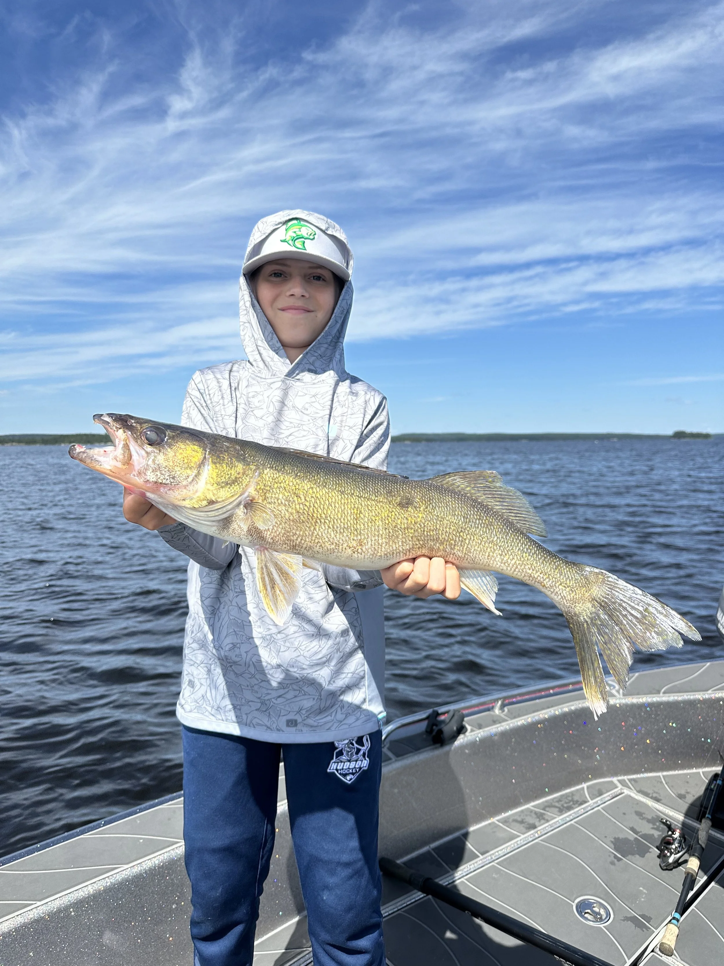 A young boy on a boat holding a large fish he caught, with a body of water and blue sky with some clouds in the background.
