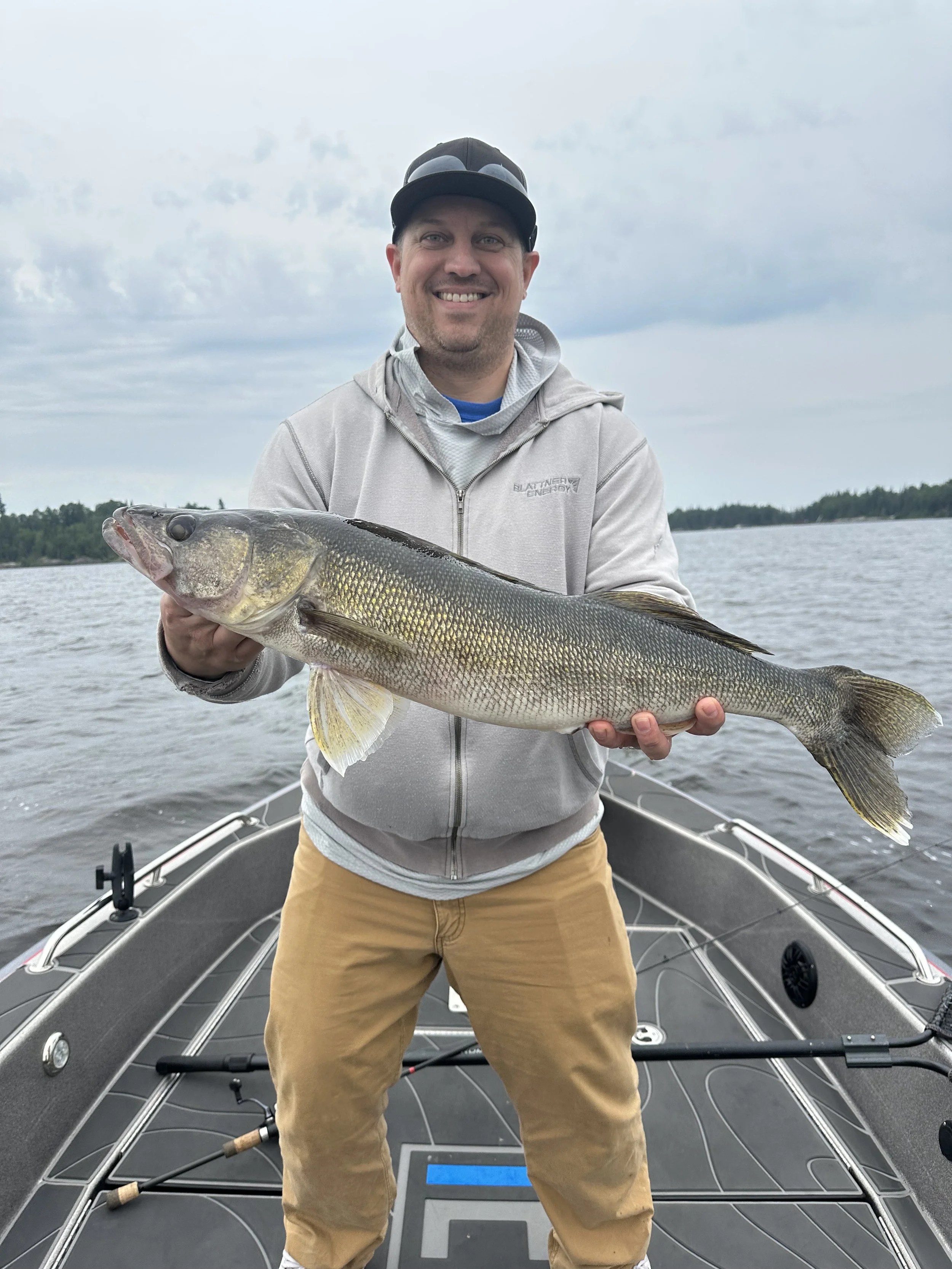 A man in a gray hoodie and tan pants holding a large fish on a boat with water and trees in the background.