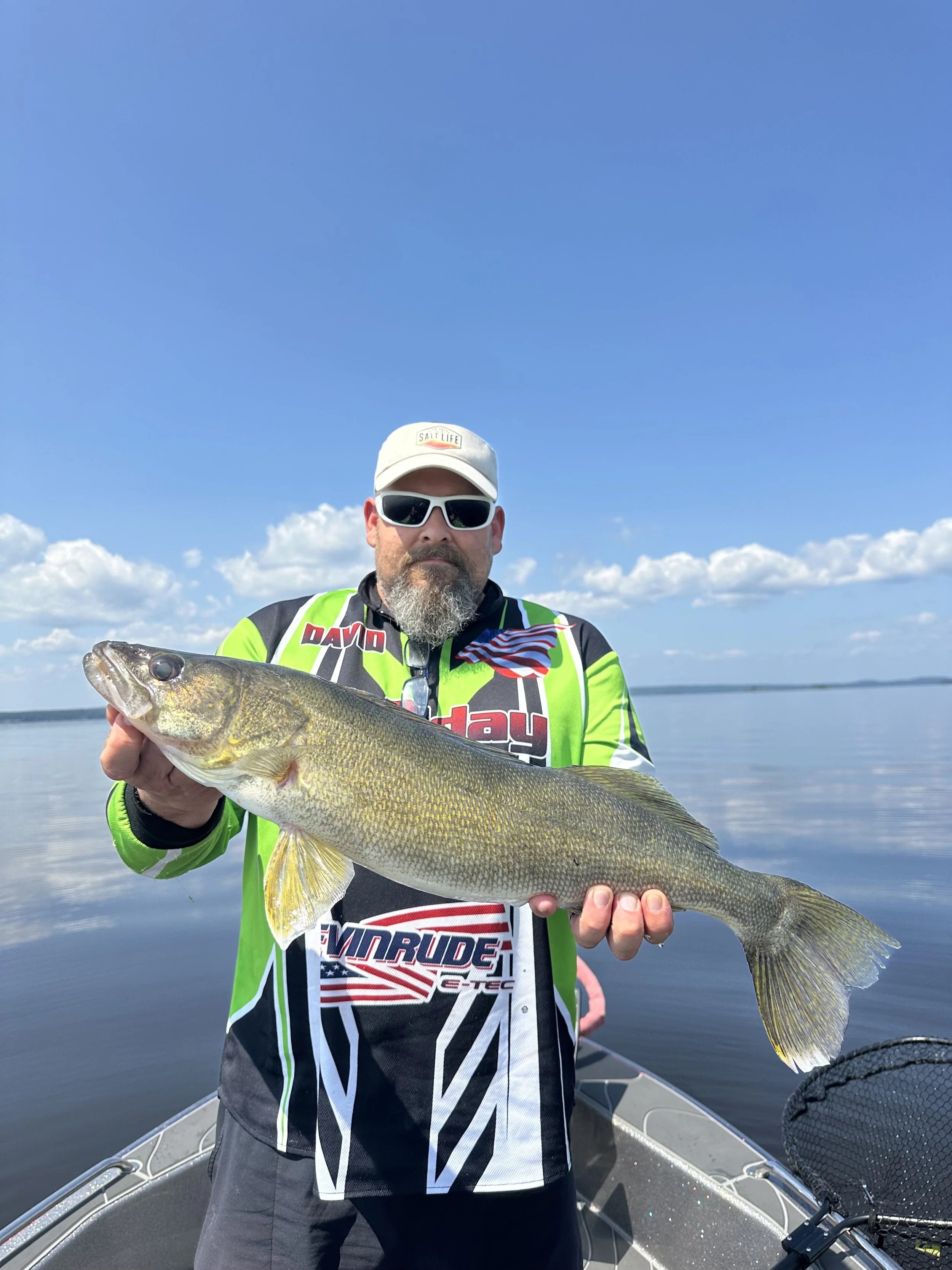 Man holding a large fish on a boat in a calm lake under a blue sky with clouds.