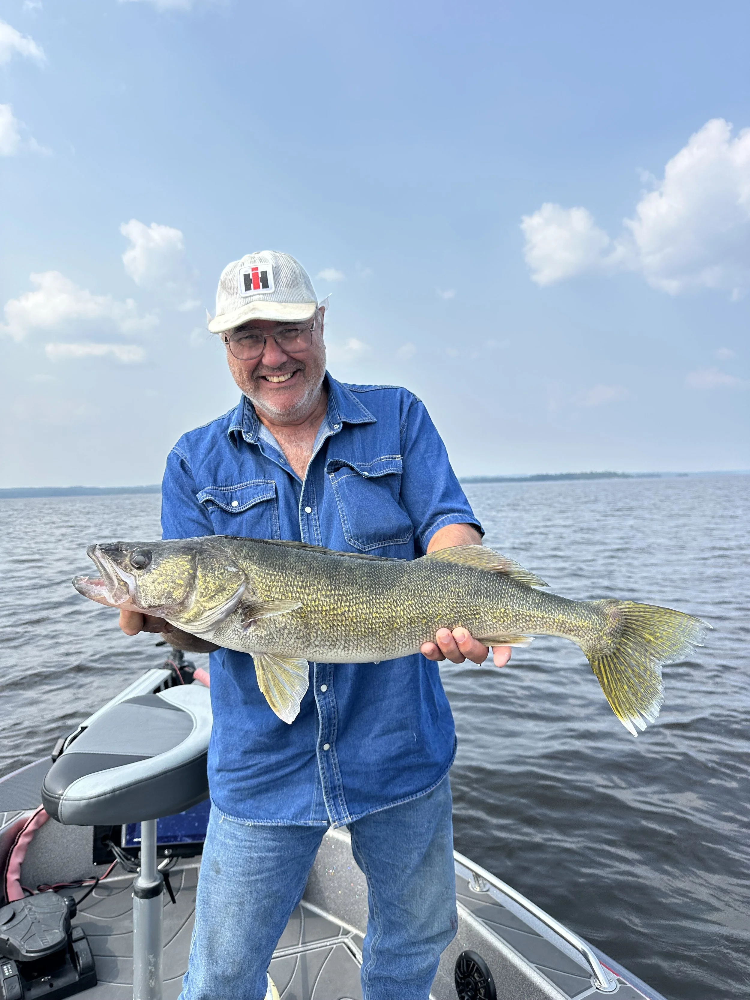 A smiling man wearing a denim shirt, glasses, and a white cap holding a large fish on a boat in the middle of a lake with a cloudy sky in the background.