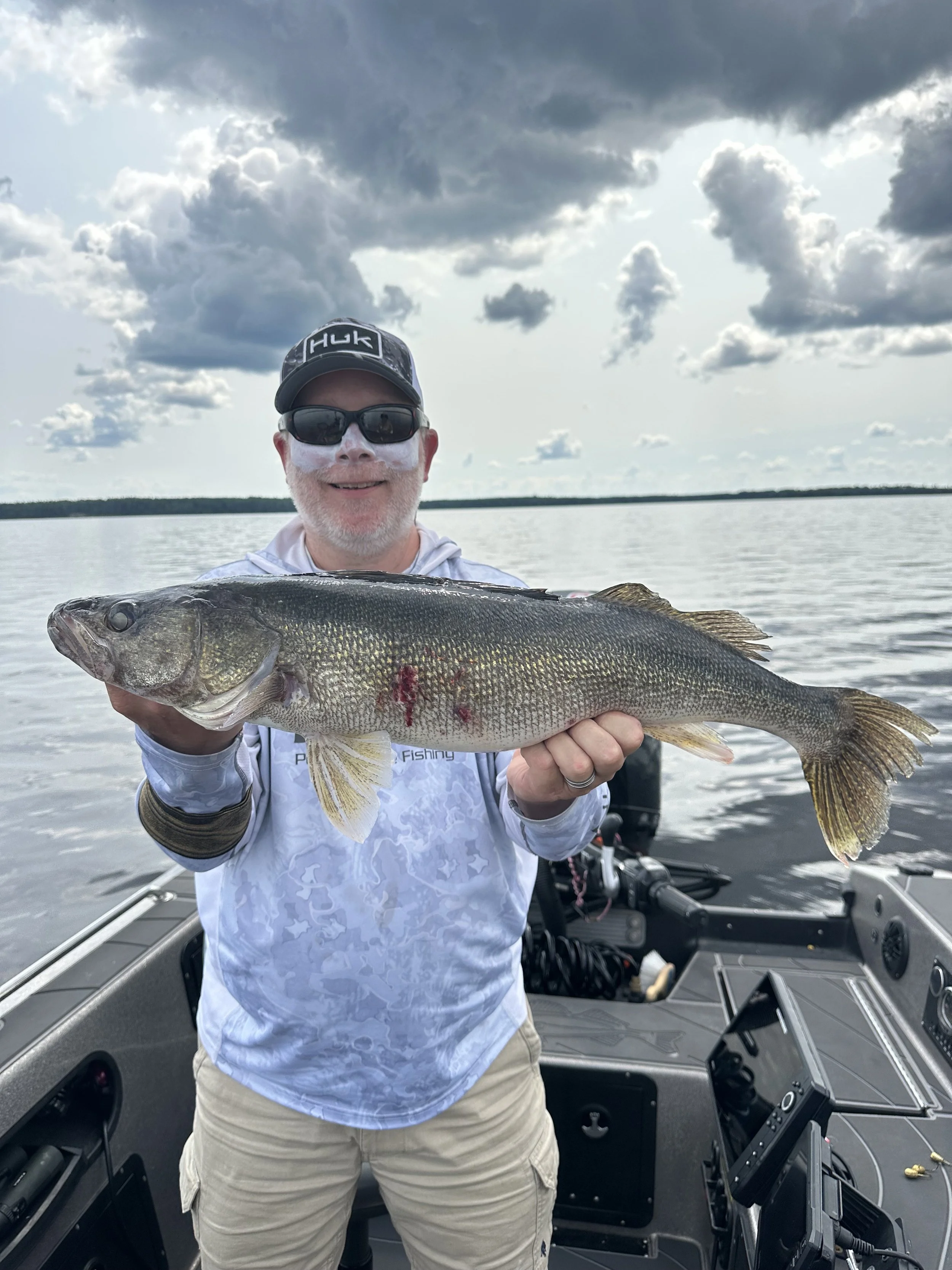 Man wearing sunglasses, a cap, and a hoodie holding a large fish on a boat in a lake.