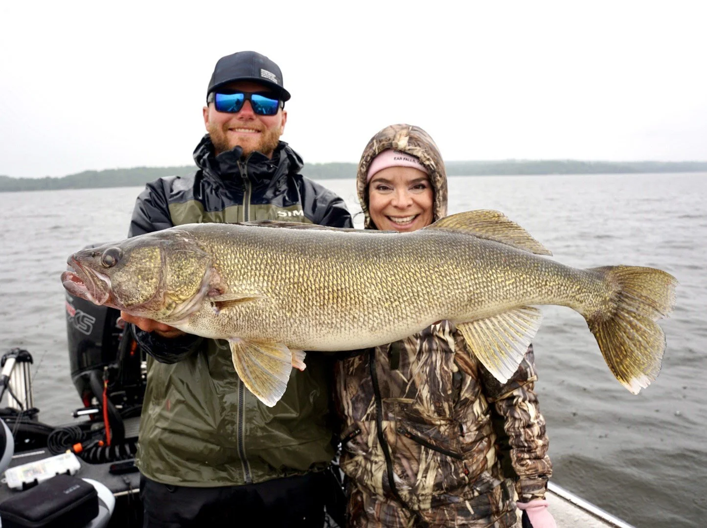 A smiling man and woman holding a large fish on a boat on the water with a distant shoreline in the background.