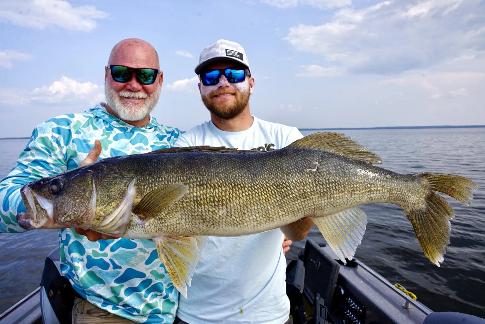 Two men on a boat holding a large fish, smiling. One man has a bald head with a white beard, wearing sunglasses and a blue patterned shirt. The other has a beard, wearing a baseball cap and sunglasses. The background shows a body of water under a par