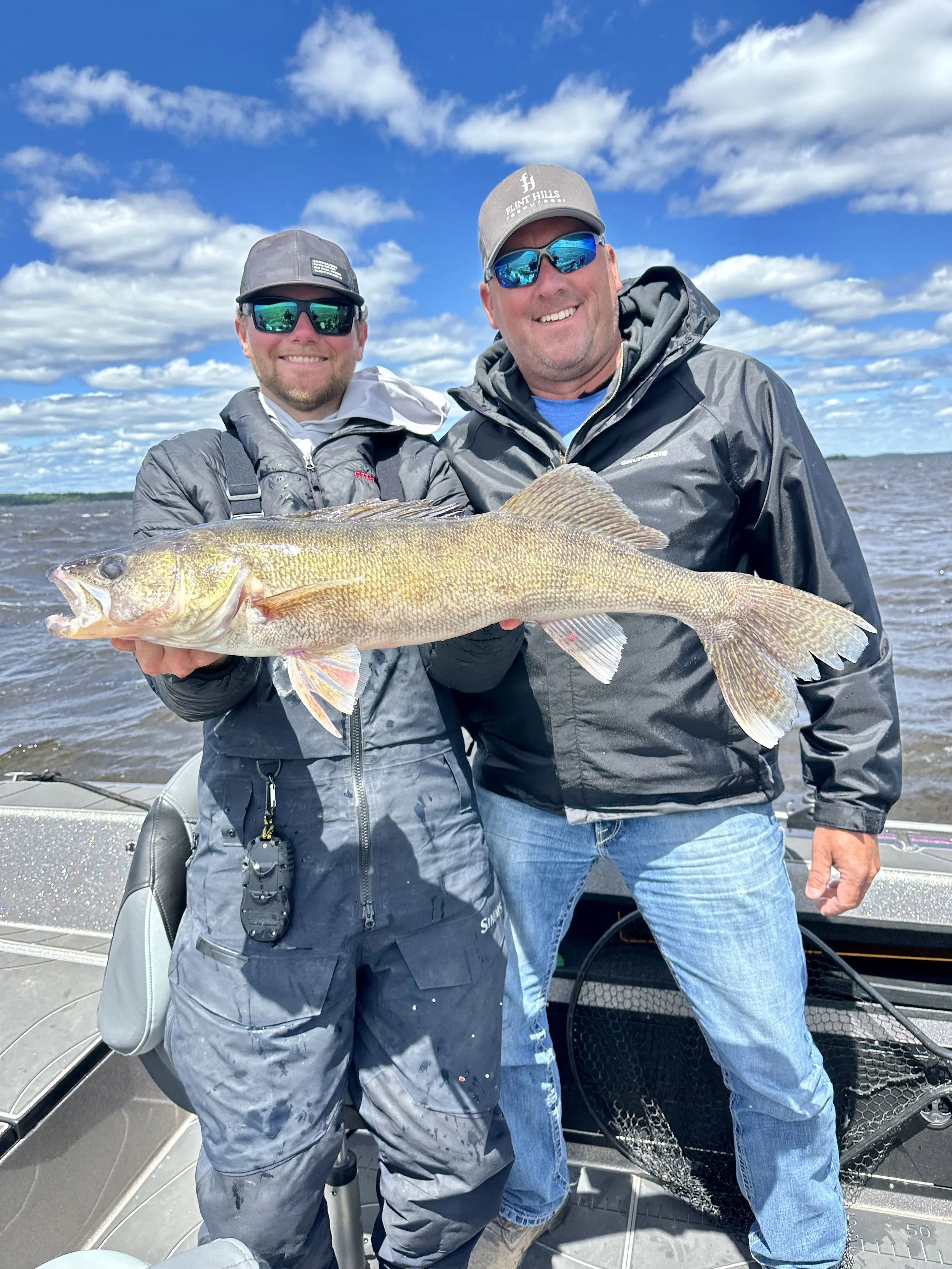 Two men in jackets and sunglasses on a boat, holding a large fish with water and sky in the background.