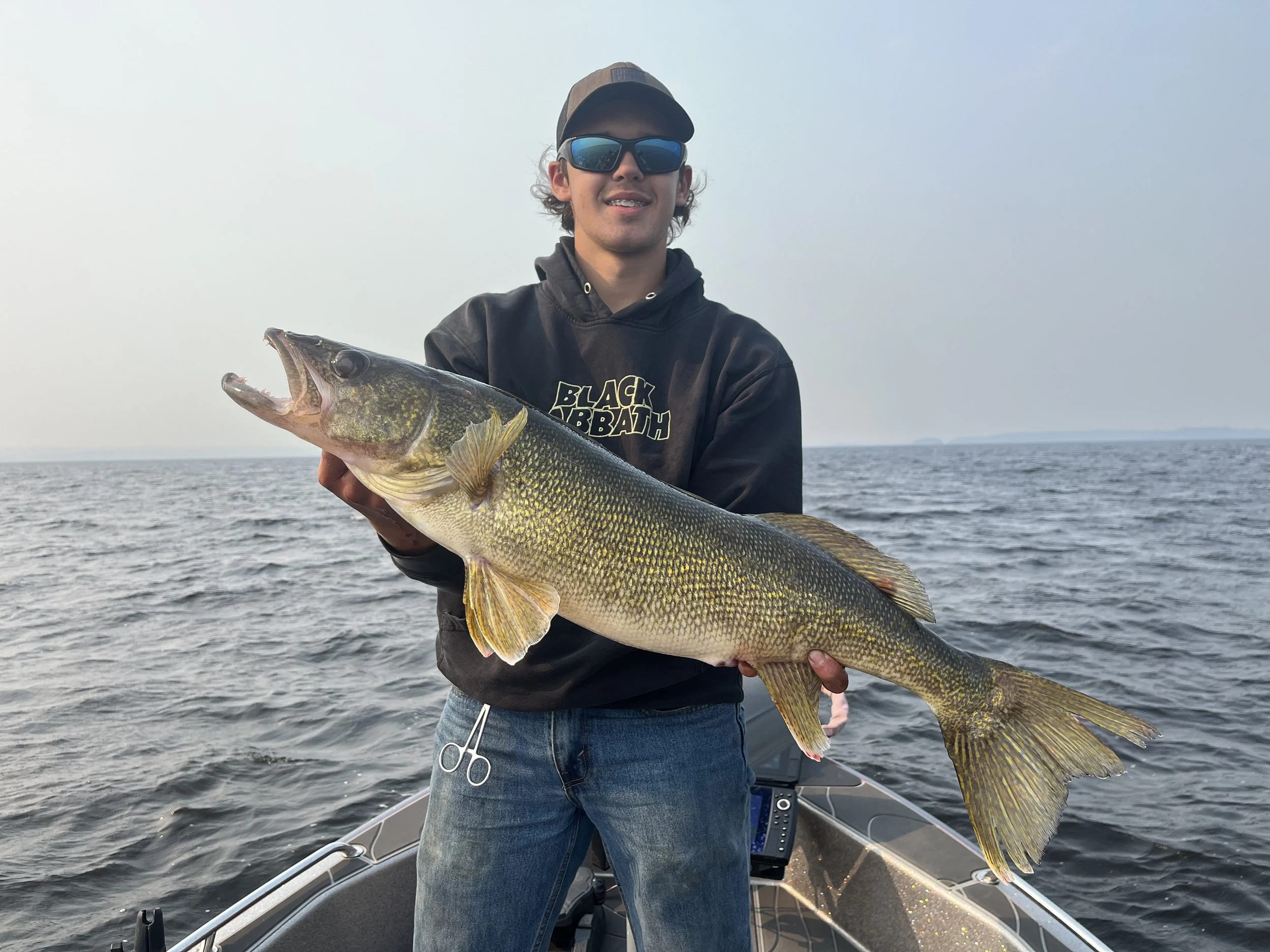 A young man wearing sunglasses, a cap, and a black hoodie standing on a boat, holding a large fish over the water.