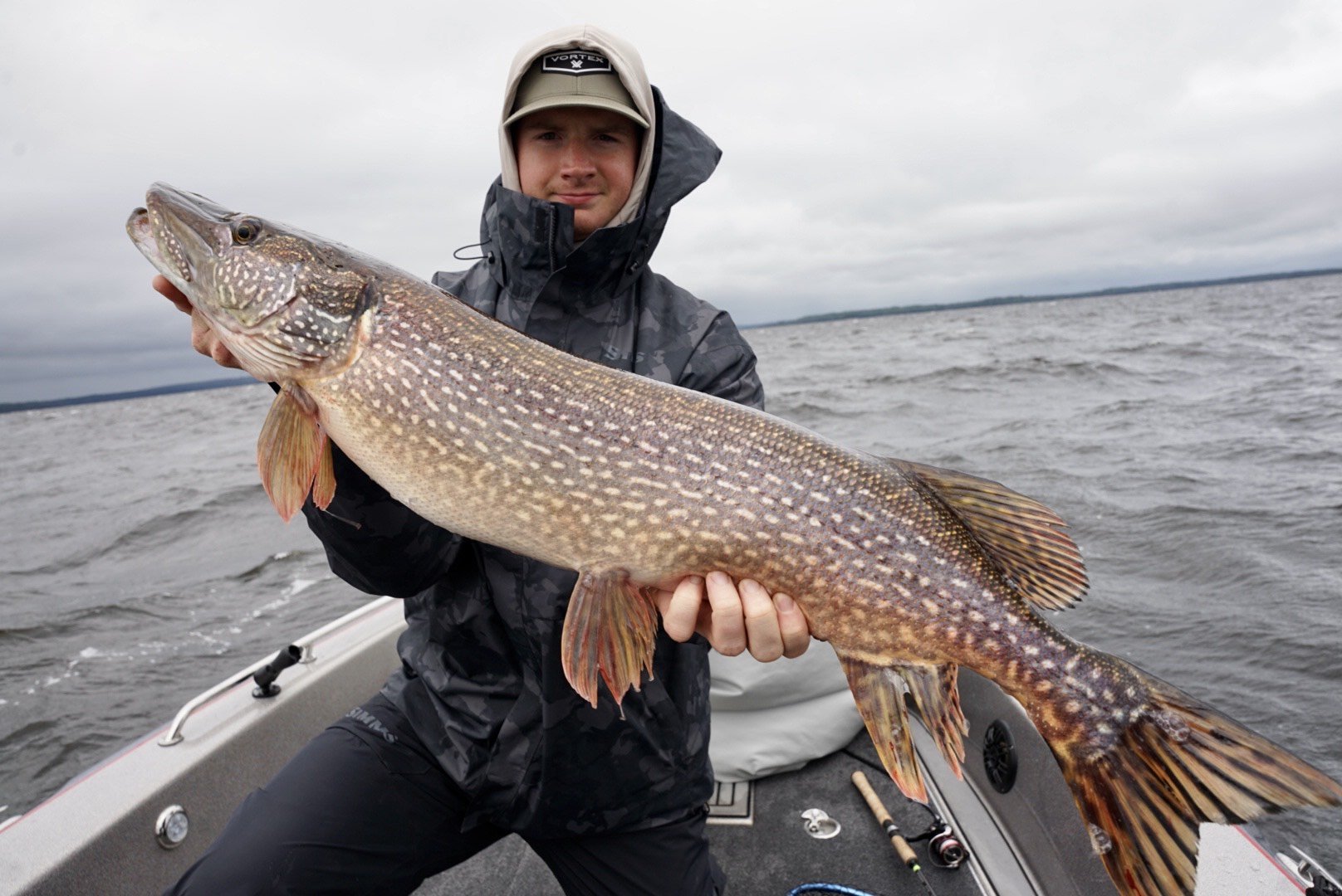 Man holding a large fish on a boat in a lake with cloudy sky.
