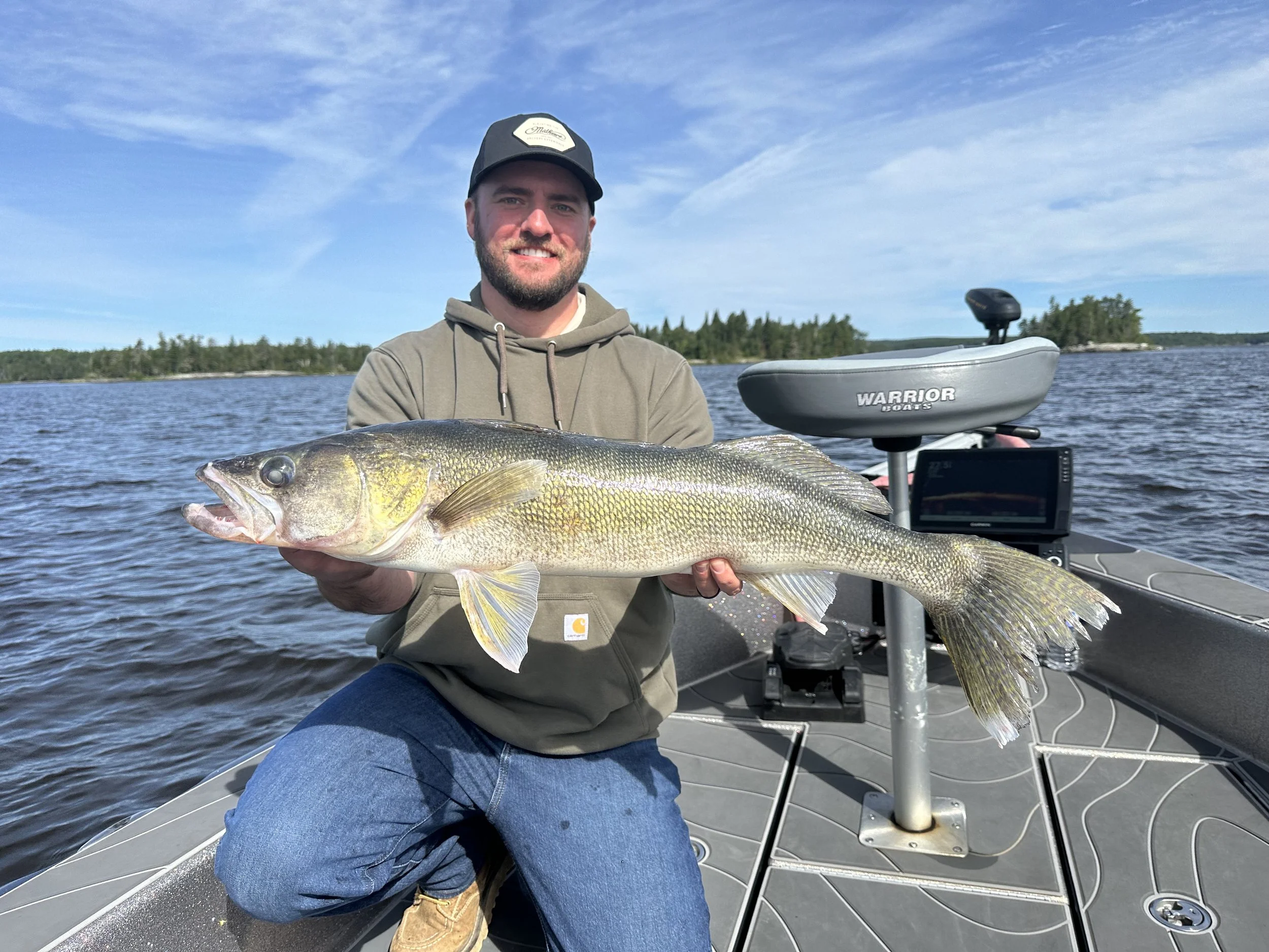 Man holding a large fish on a boat in a lake with trees in the background under a partly cloudy sky.