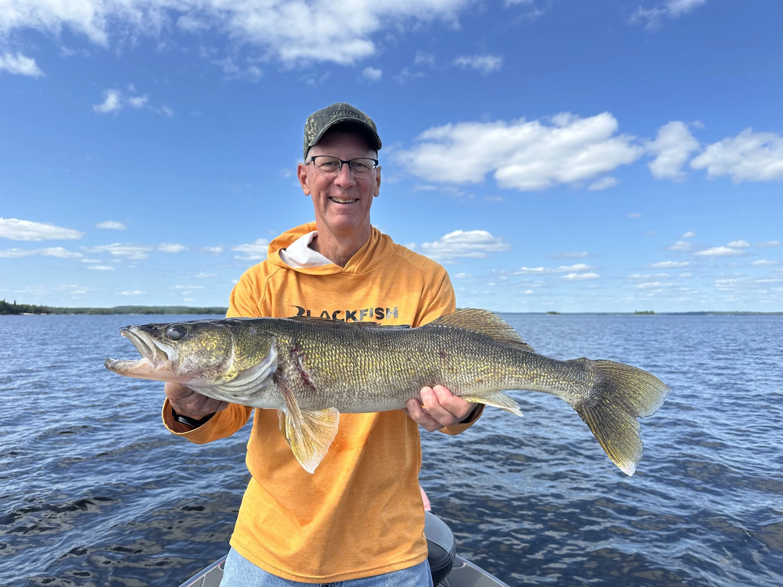 A man with glasses and a cap holding a large fish on a boat in a lake under a blue sky with some clouds.