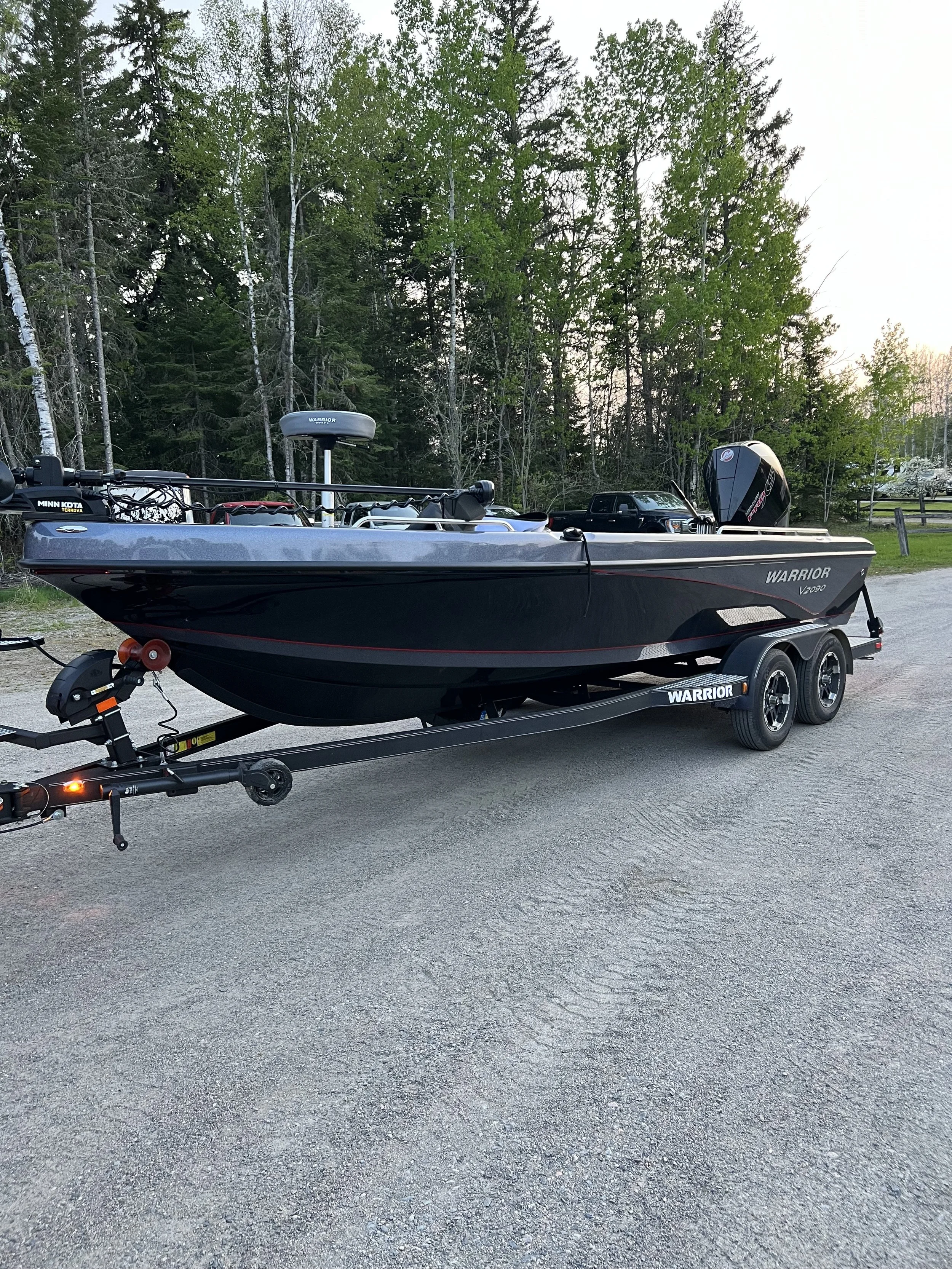 A black Warrior V200 boat on a trailer, parked on a gravel surface near a wooded area with green trees, and a black truck in the background.