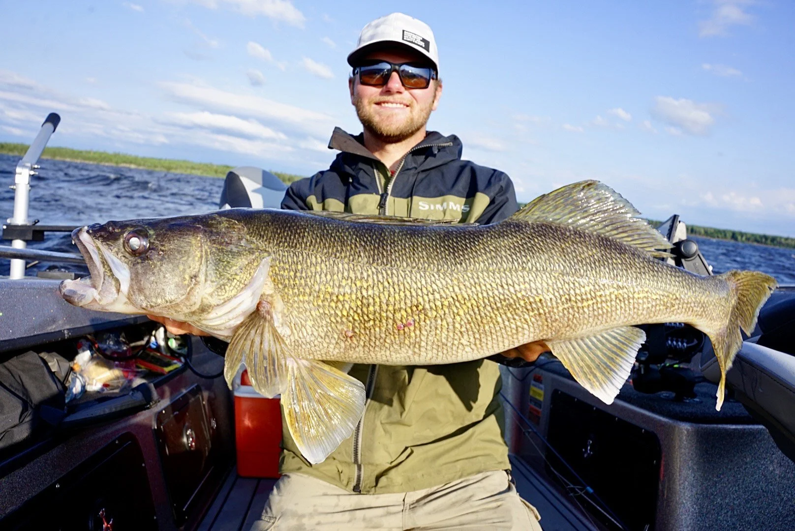 Man in sunglasses and a cap holding a large fish on a boat in a lake with blue sky and clouds.