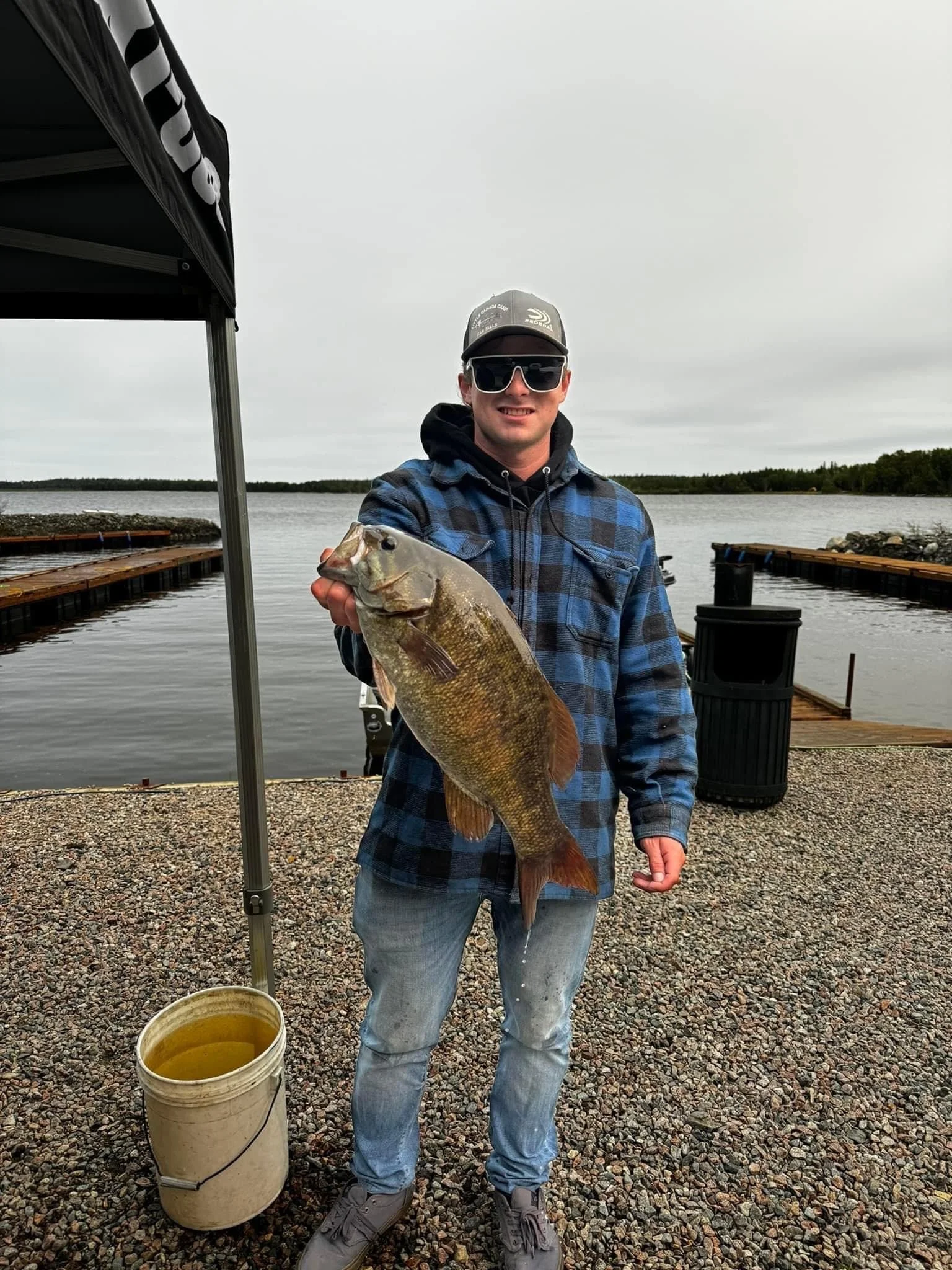 A man in a plaid jacket, jeans, and sunglasses holding a large fish by a body of water with a dock in the background.