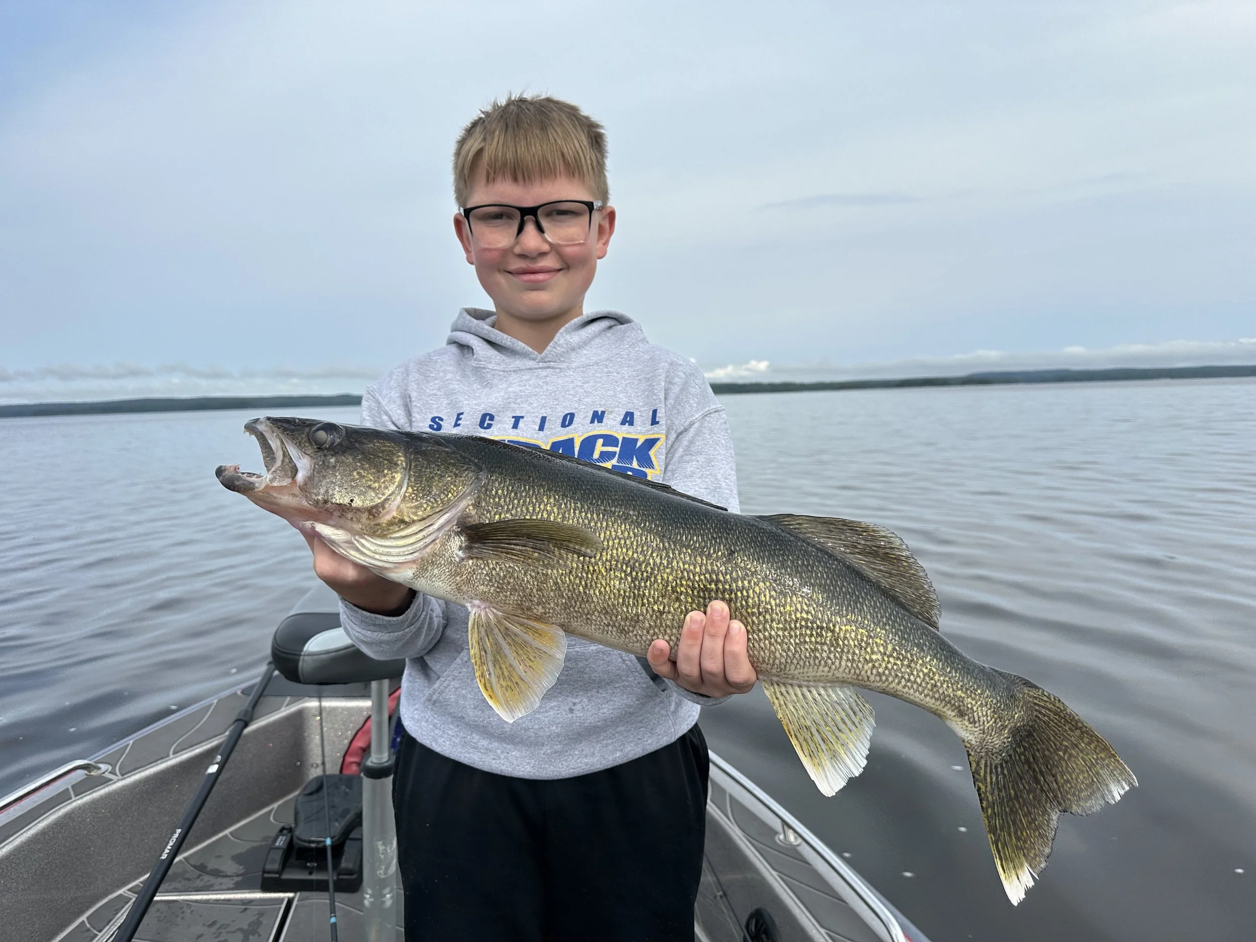 A boy in a gray hoodie and glasses smiling while holding a large fish on a boat in a large body of water.