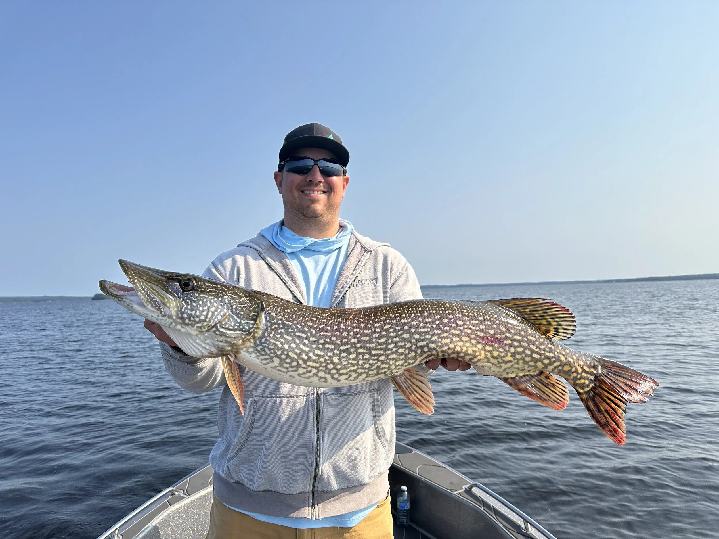 A man wearing sunglasses and a hoodie holding a large colorful fish on a boat in the water, with a clear sky and distant shoreline in the background.