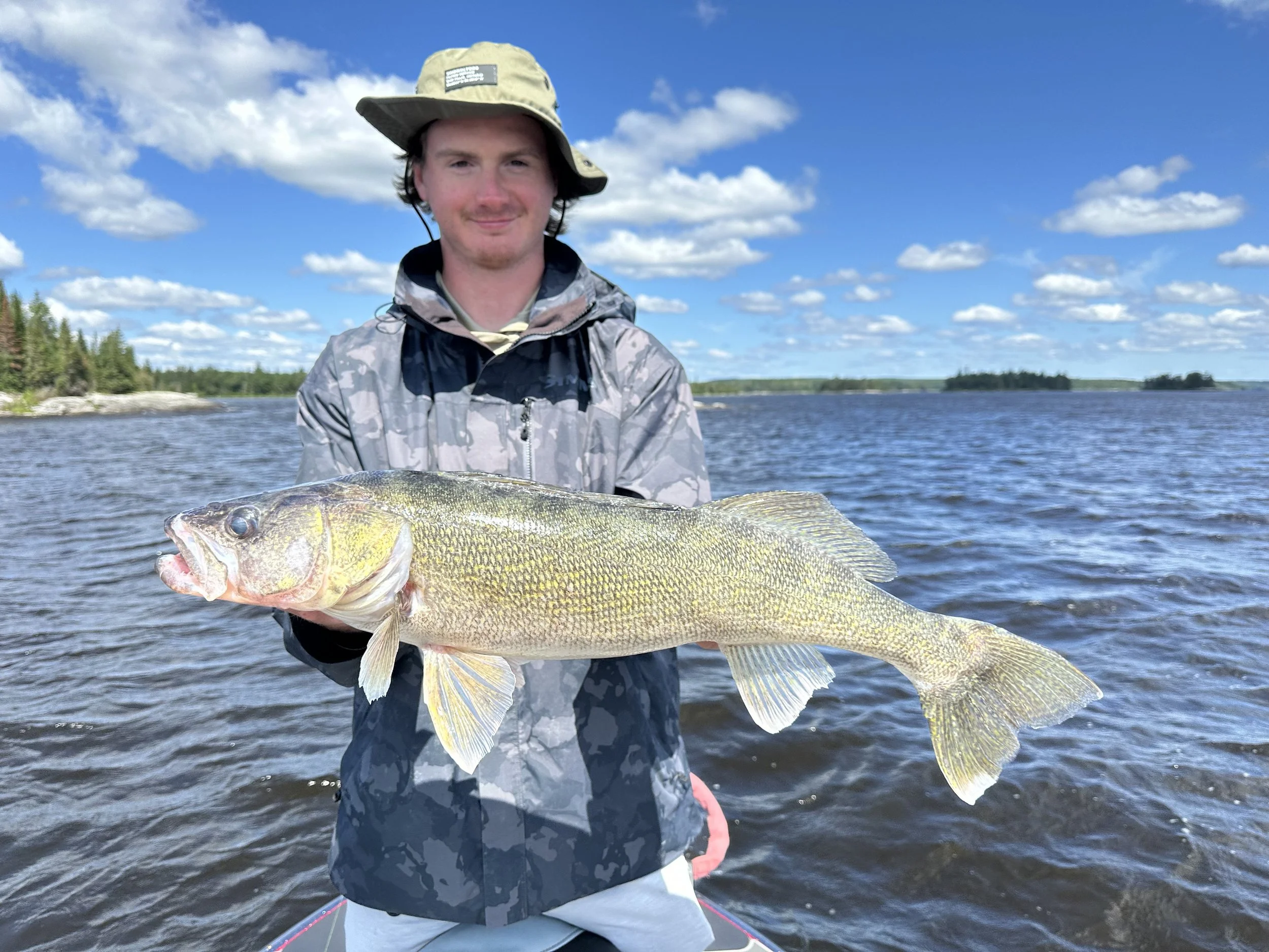 A man in outdoor clothing holding a large fish on a body of water with a partly cloudy sky in the background.