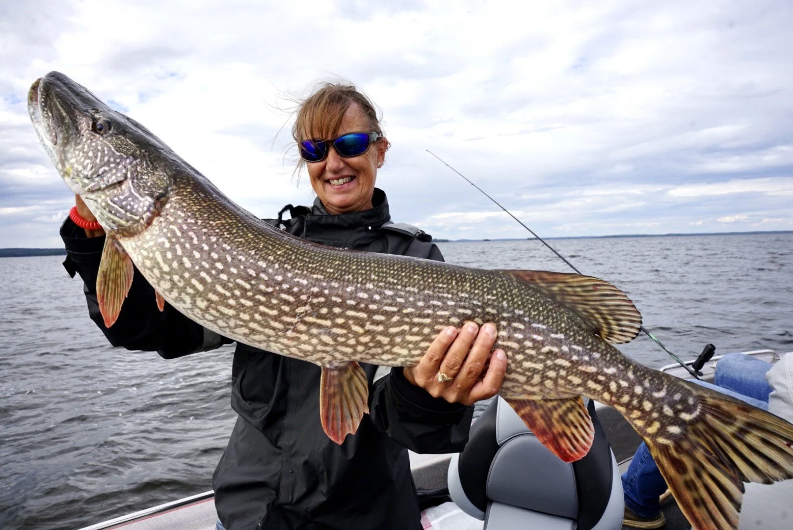 Woman holding a large fish on a boat, with water and cloudy sky in the background.