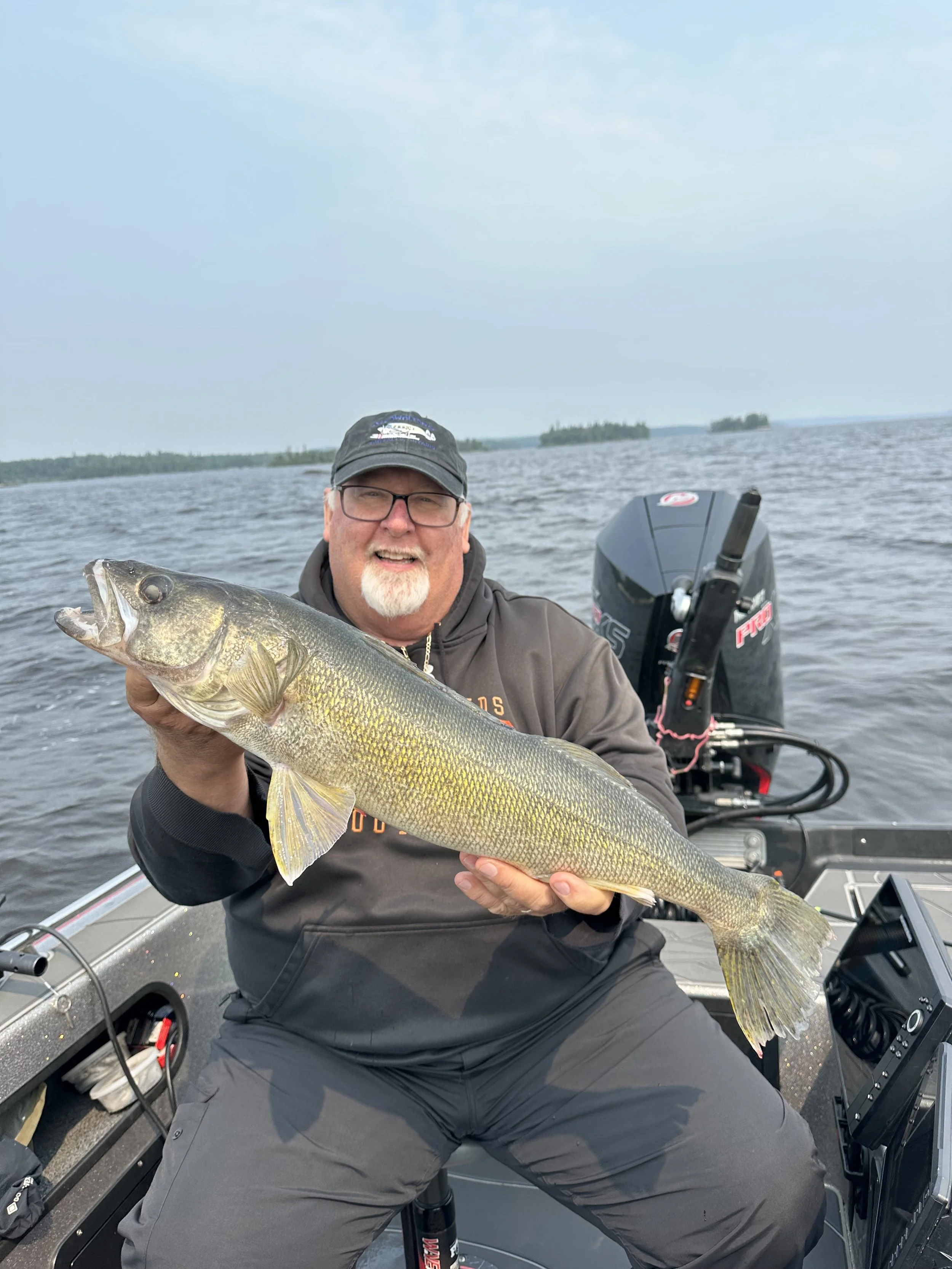 Man in a boat holding a large fish, a walleye, with a cloudy sky and water in the background.