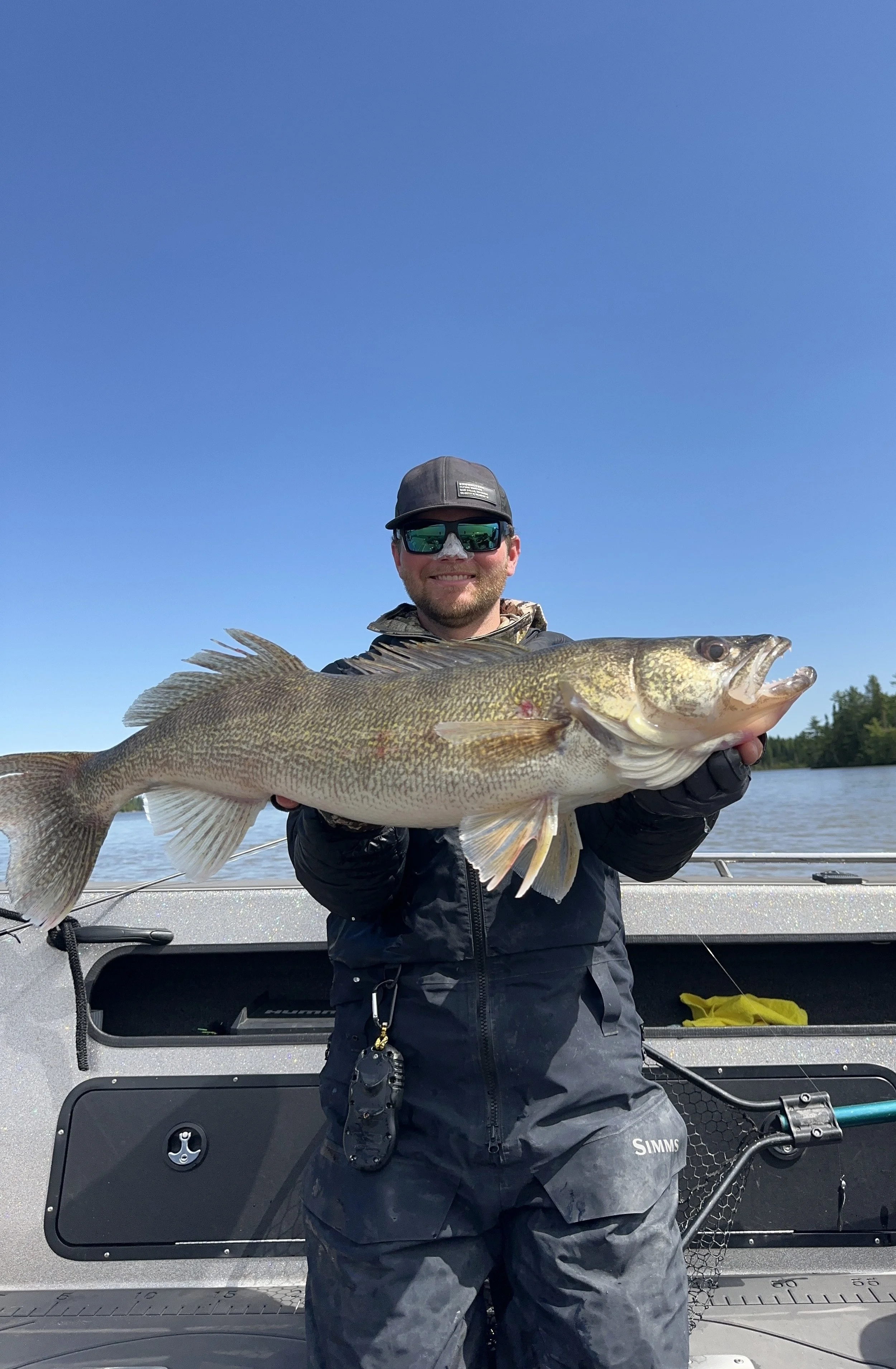 Man in sunglasses and dark fishing gear holding a large fish on a boat with a clear blue sky in the background.