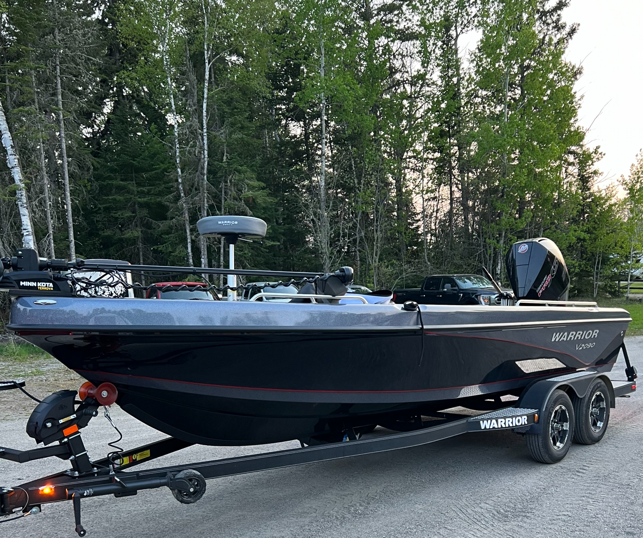 A black Warren fish boat on a trailer parked on a gravel surface with trees and vehicles in the background.