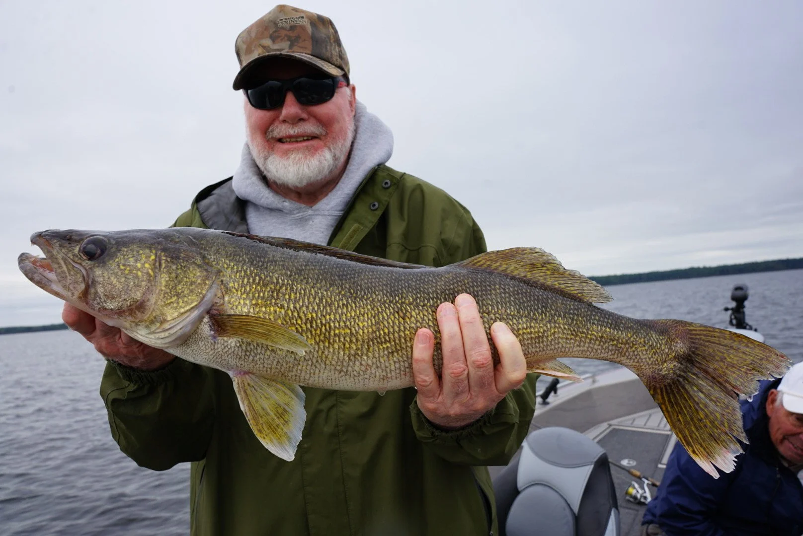 A smiling man with sunglasses, a camouflage hat, and a green jacket holds a large fish over a body of water, with another man sitting nearby in a boat.