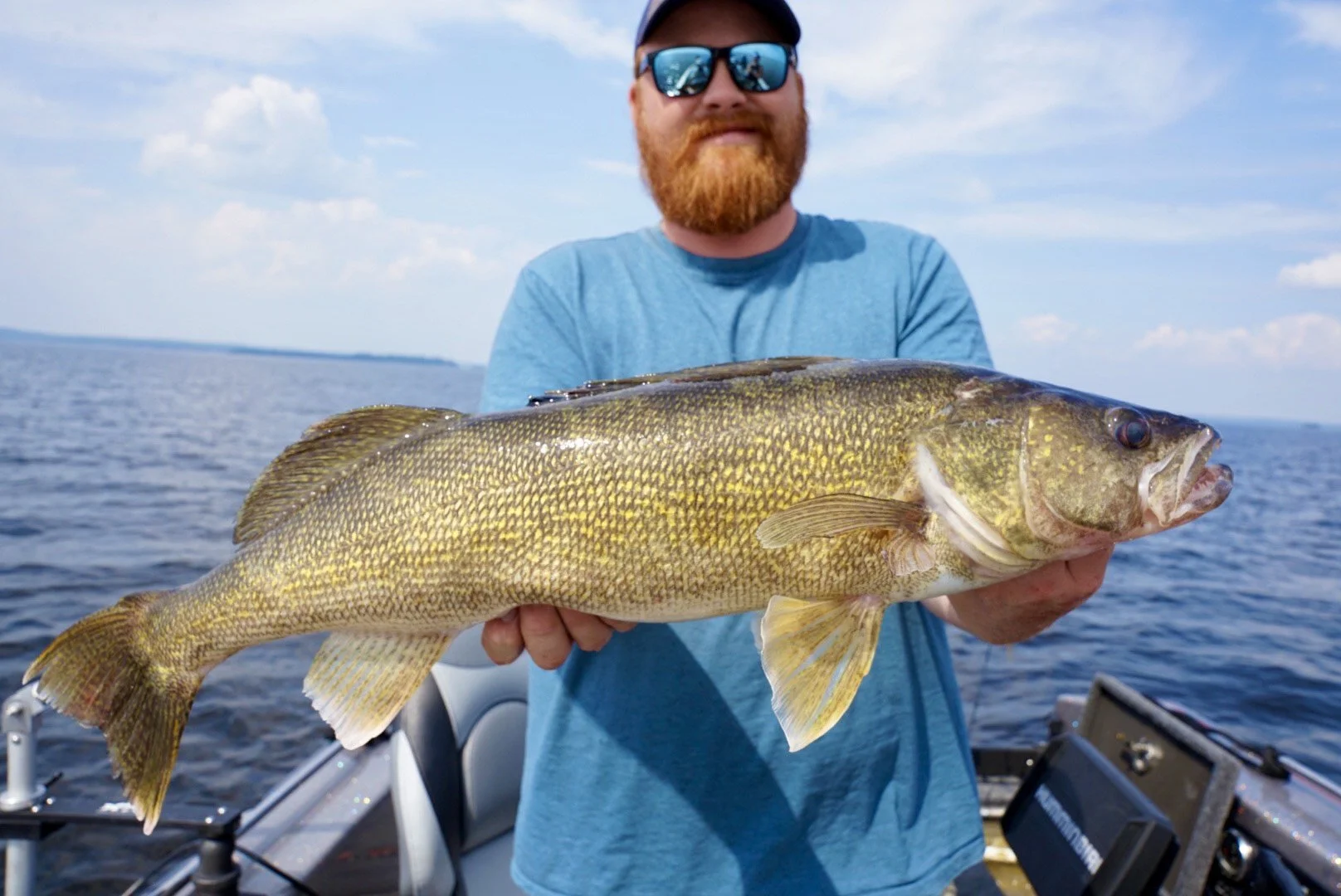 A man with a red beard and sunglasses holding a large fish on a boat in the water.