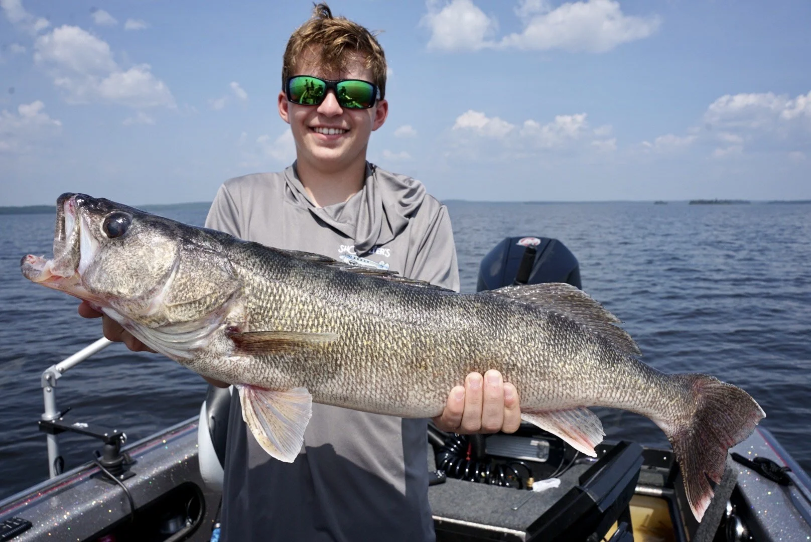A smiling young man with sunglasses holding a large fish on a boat in the water during daytime.