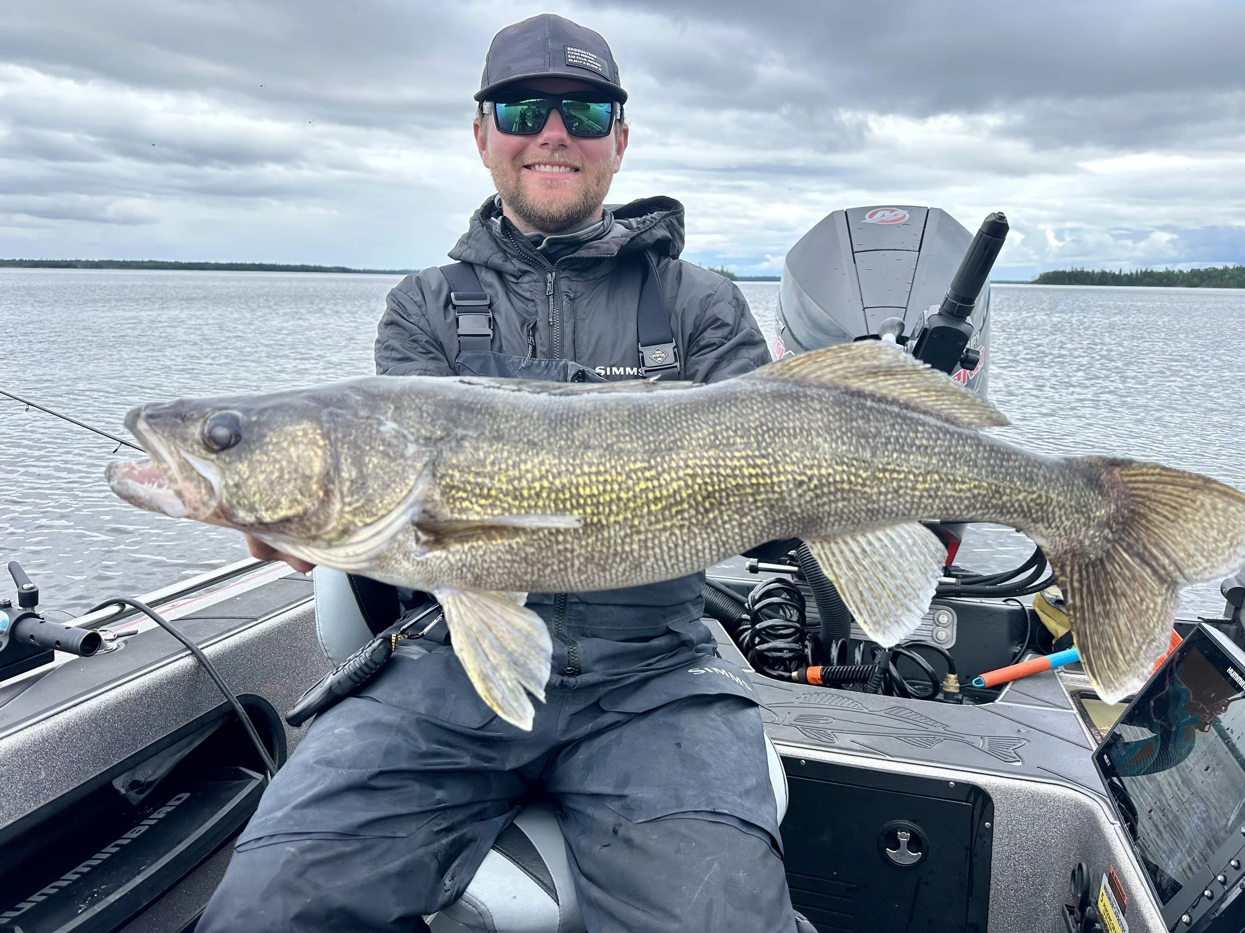 Man smiling on a boat holding a large fish with a lake and cloudy sky in the background.