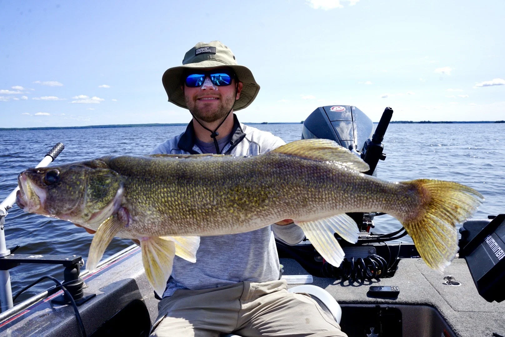 Man on a boat holding a large fish, lake in background, wearing hat and sunglasses.