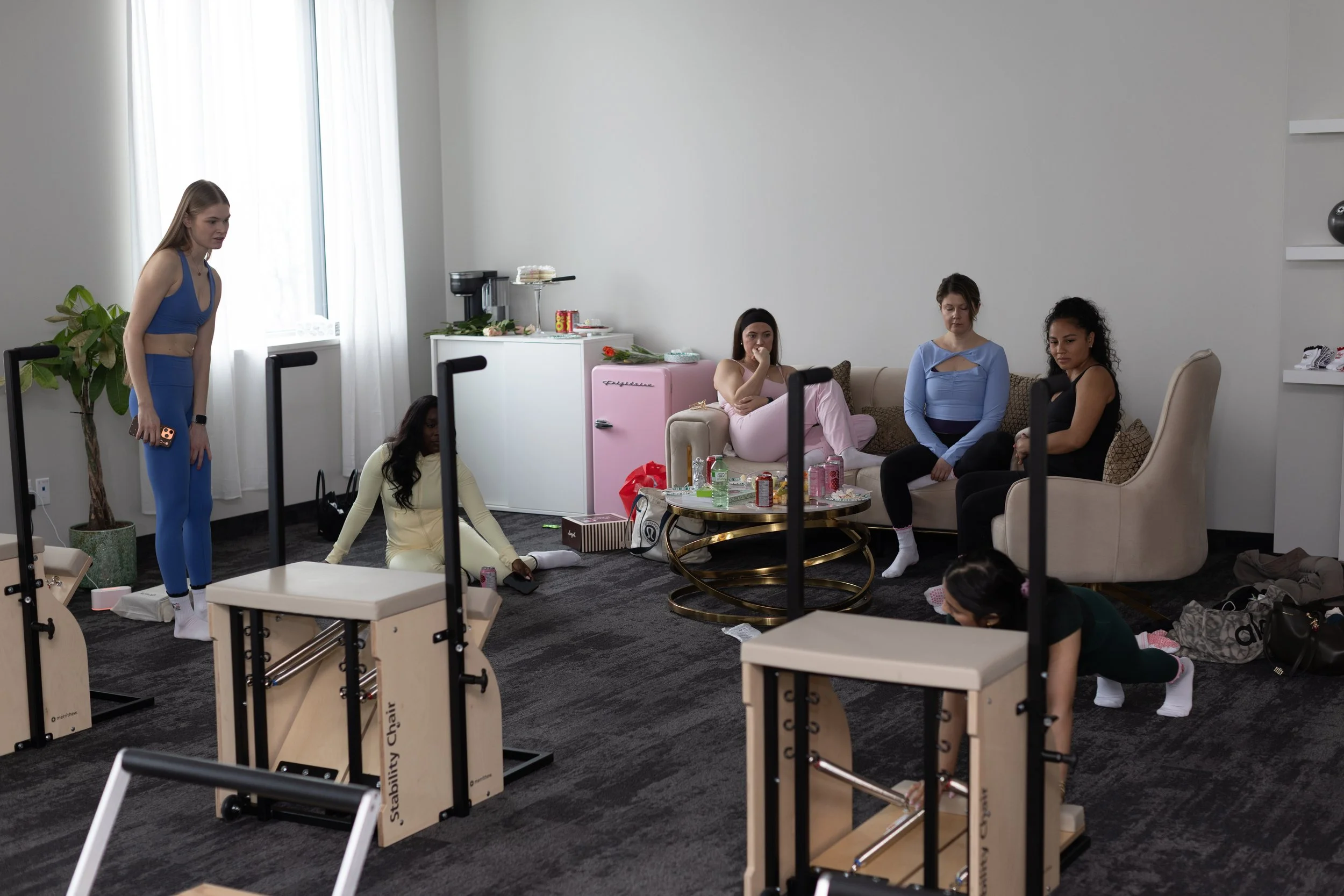 Group of women exercising and stretching in a fitness room with Pilates equipment and a small group of spectators on the sofa