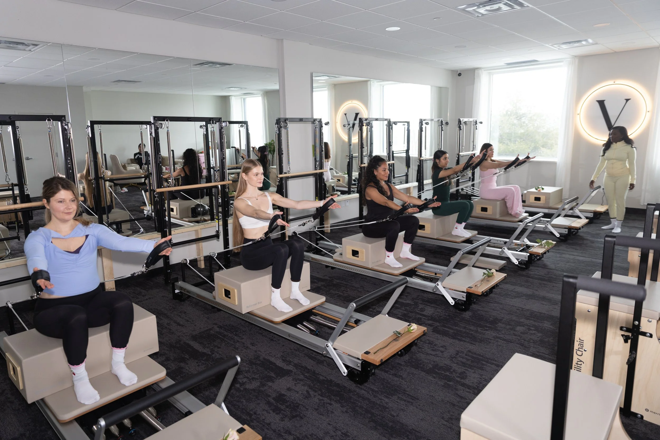 Women using Pilates reformer machines in a fitness studio with large mirrors and bright natural light.