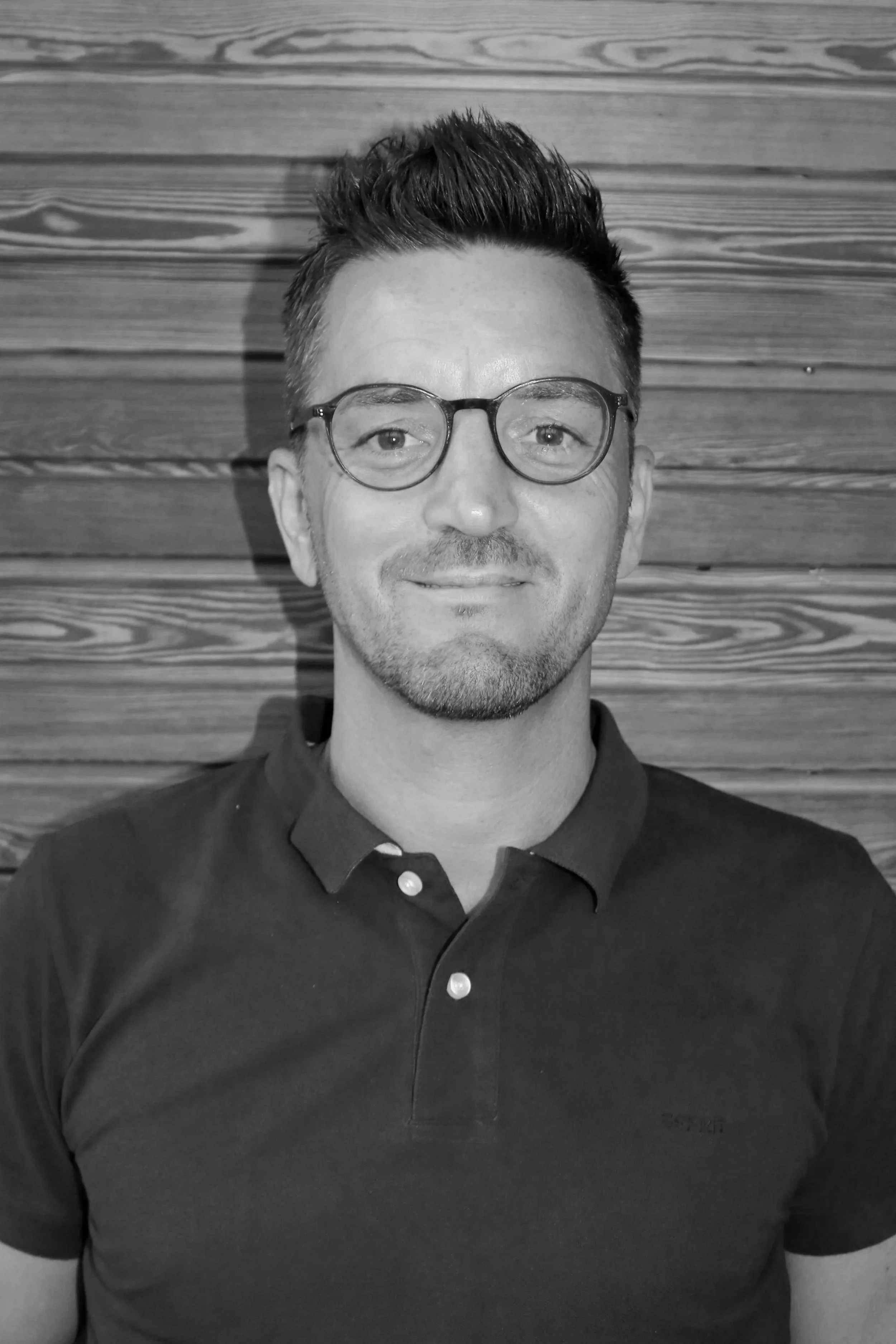 Schwarz-Weiß-Fotografie eines Mannes mit Kurzhaarfrisur, Brille und gepflegtem Bart, trägt ein Polo-Hemd, vor einer Holz-Wand.