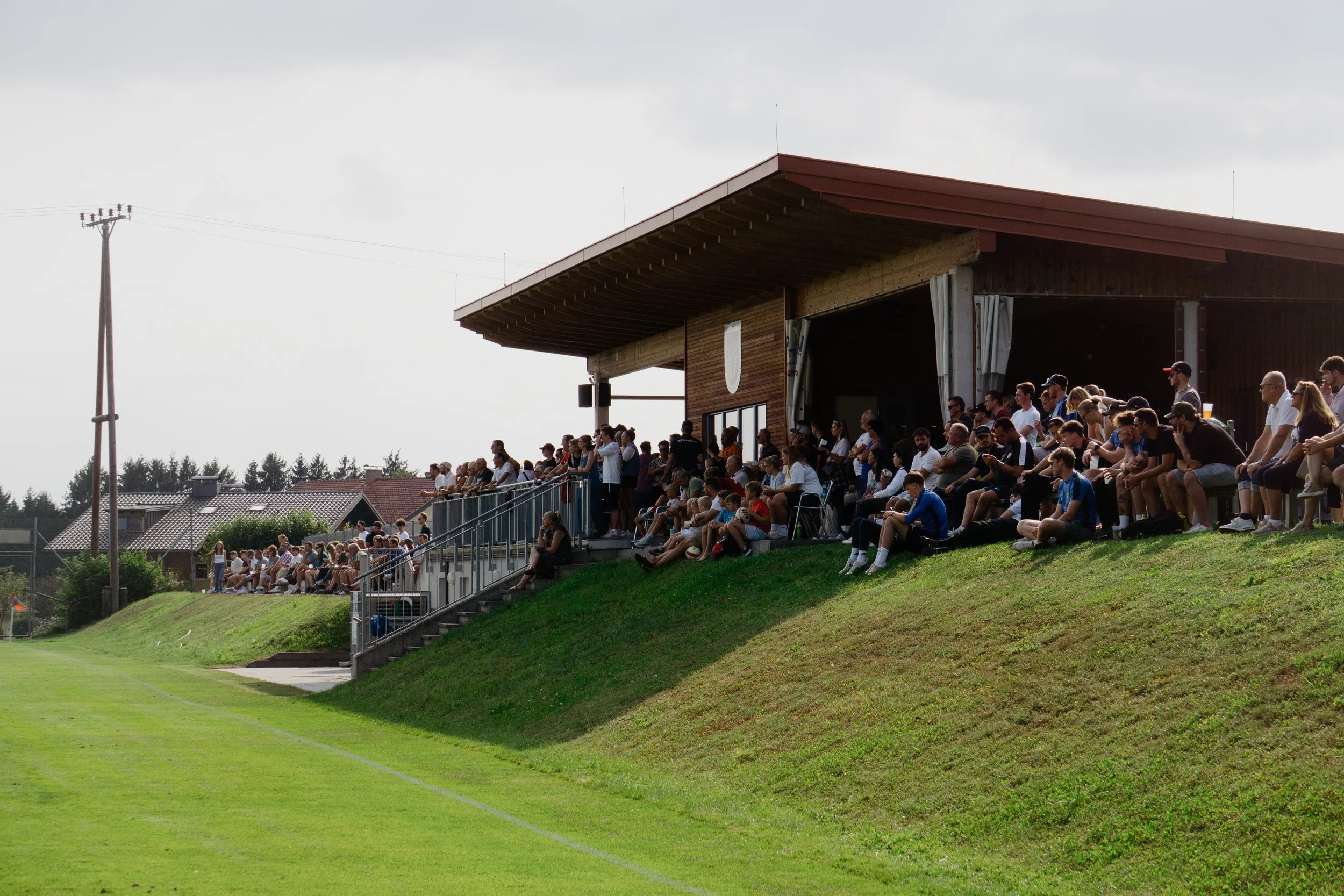 Menschen sitzen auf einer Tribüne bei einem Fußballspiel im Freien, einige im Schatten eines Holzdachüberbaus.
