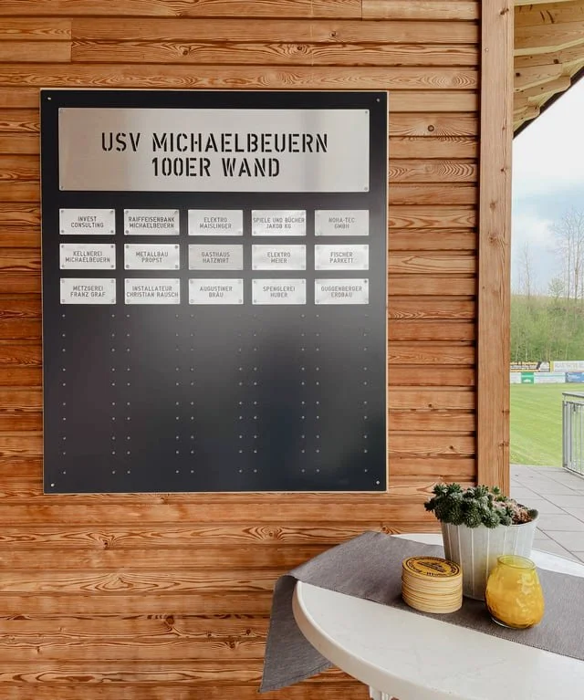 Ein schwarzes schwarzes Whiteboard mit der Aufschrift "USV Michaelbeuern 100er Wand" und mehreren Namen und Tätigkeiten, an einer Holzverkleidung im Freien, neben einem Tisch mit Pflanzen und Dekorationen.