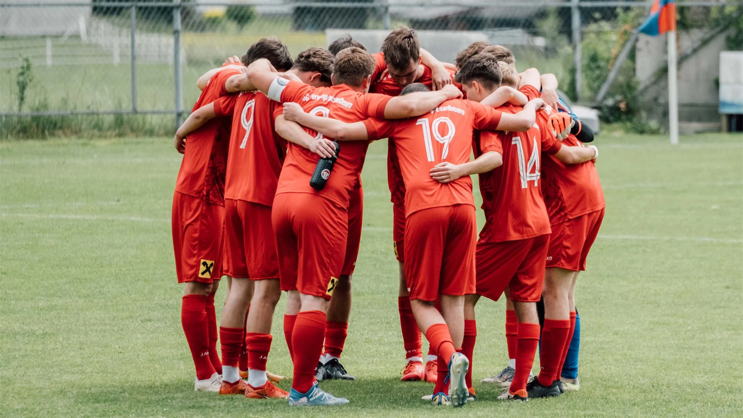 Fußballteam in roter Uniform versammelt sich in einem Kreis auf dem Spielfeld bei einem Mannschaftszeichen.