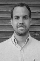 Schwarz-weiß-Fotografie eines Mannes, der vor einer Holzlattenwand steht und lächelt.