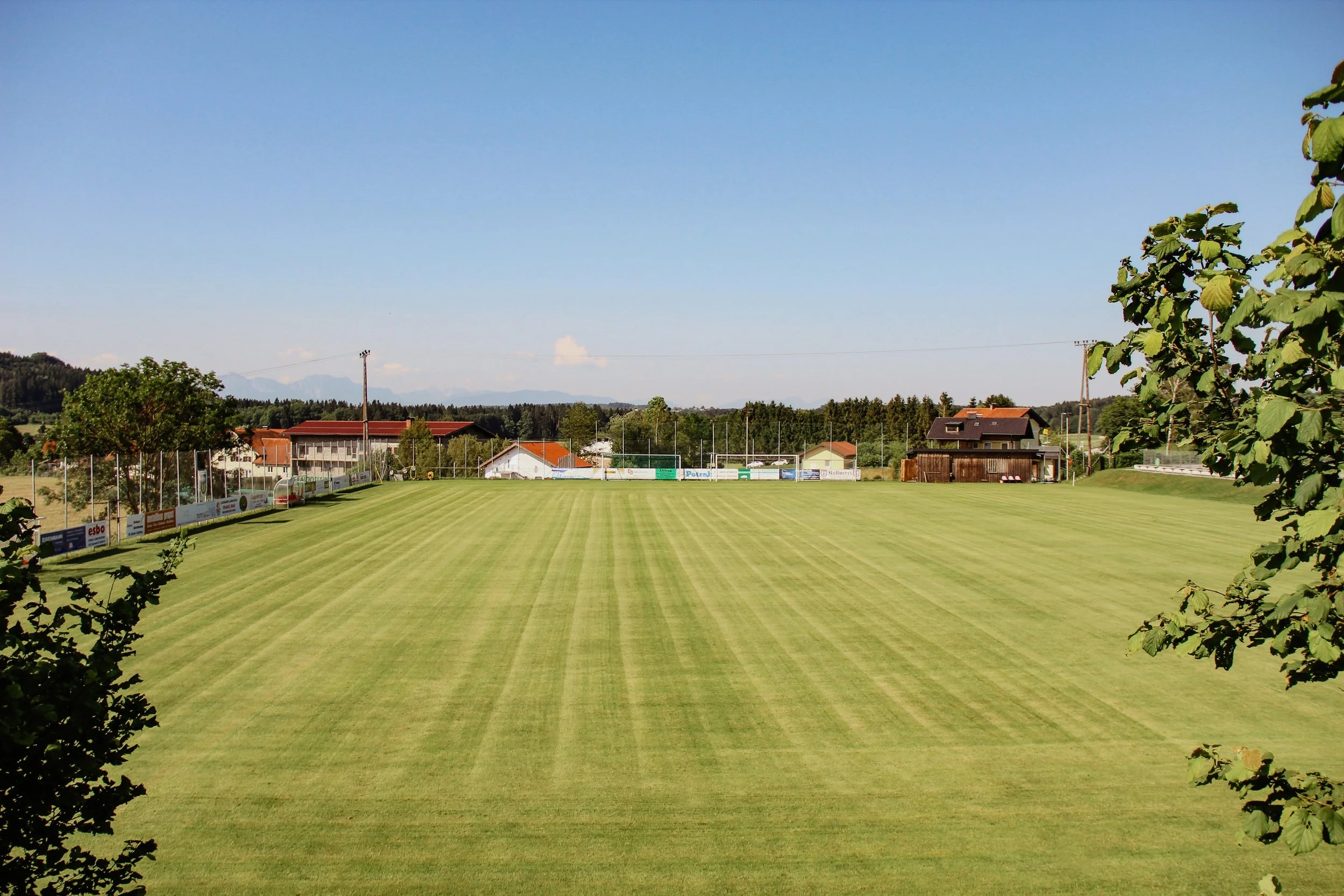 Ein ausgestatteter Fußballplatz in einer ländlichen Gegend mit Häusern im Hintergrund, umgeben von Bäumen und einem klaren blauen Himmel.