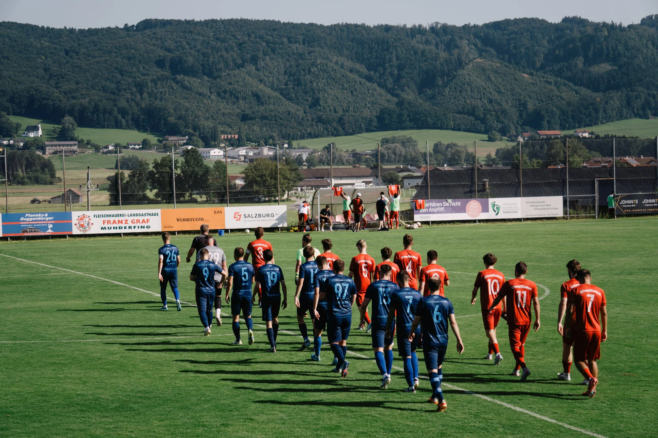 Fußballspieler auf dem Spielfeld, die Mannschaften in blauen und roten Trikots, mit einem Bergland im Hintergrund.