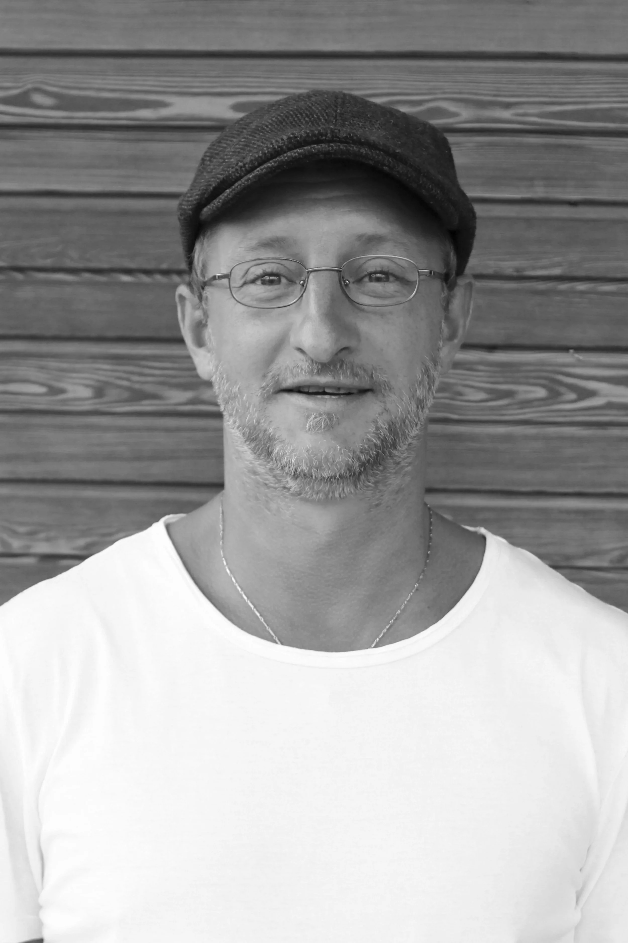 Schwarz-weiß-Foto eines Mannes mit Brille, grauem Bart und Mütze, vor einer Holzwand