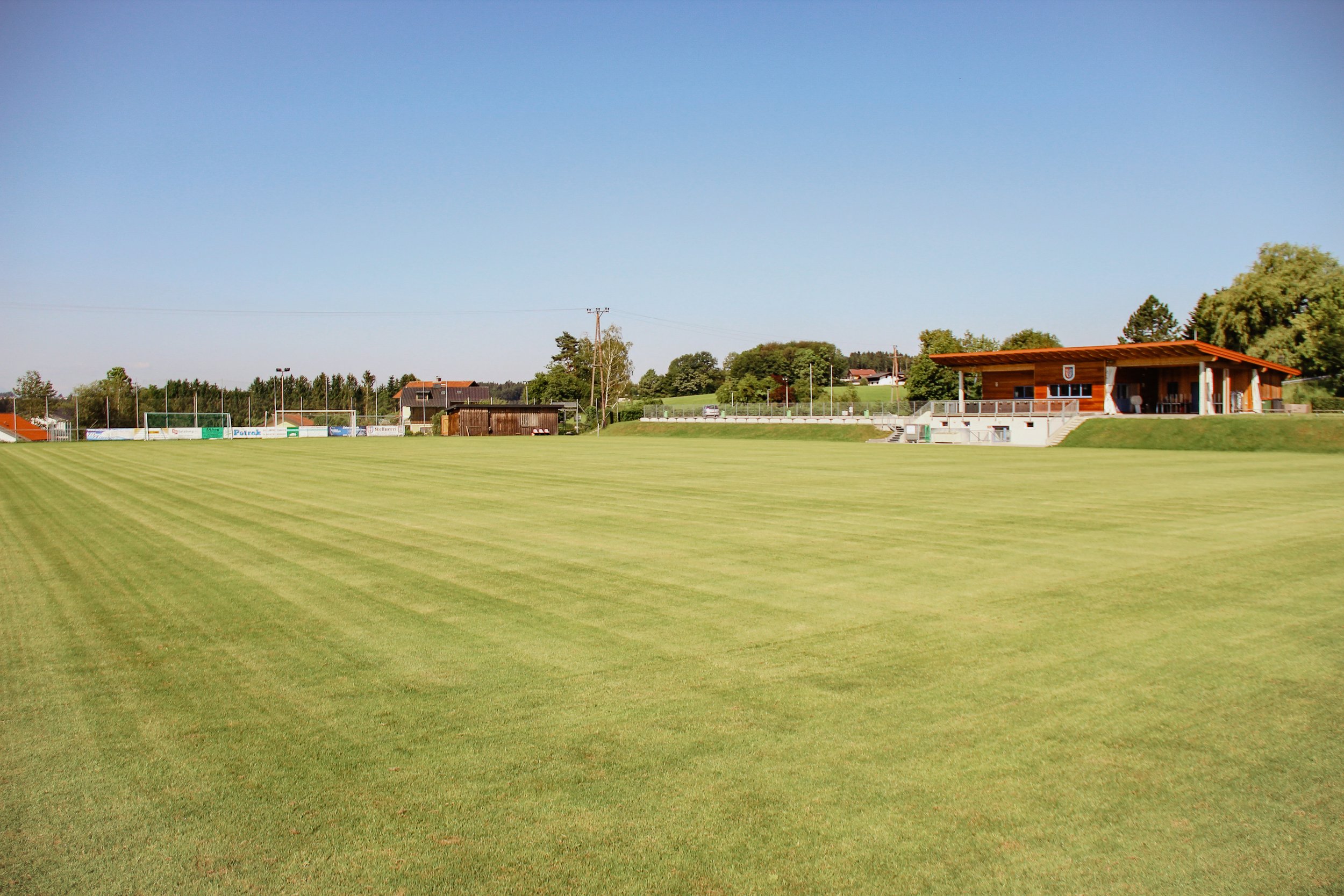 Leeres Fußballfeld mit grünem Rasen, einem kleinen Gebäude auf der rechten Seite und Bäumen im Hintergrund under blauer Himmel.