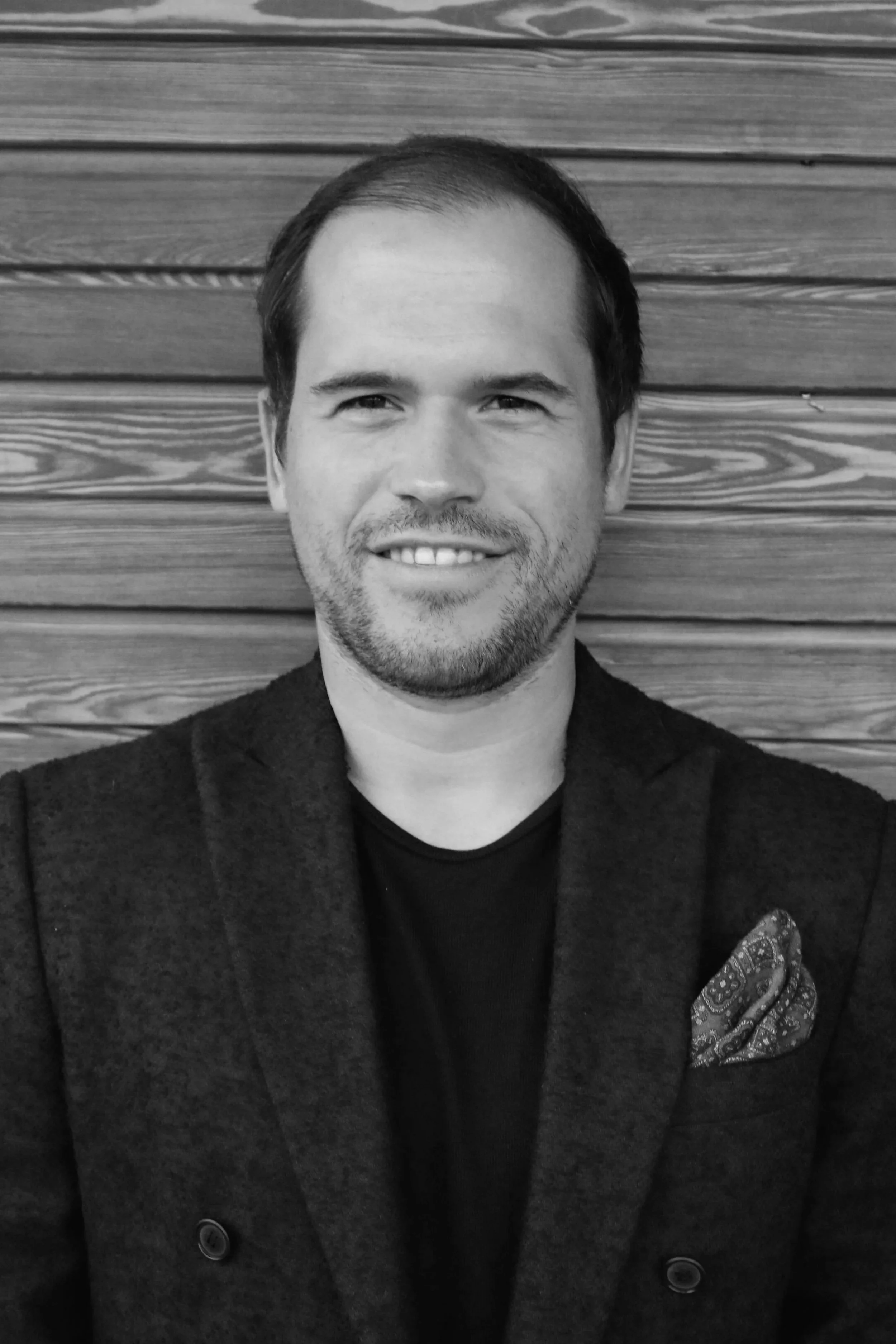 Ein lächelnder Mann mit dunklem Haar und Bart vor einer Holzverkleidung.