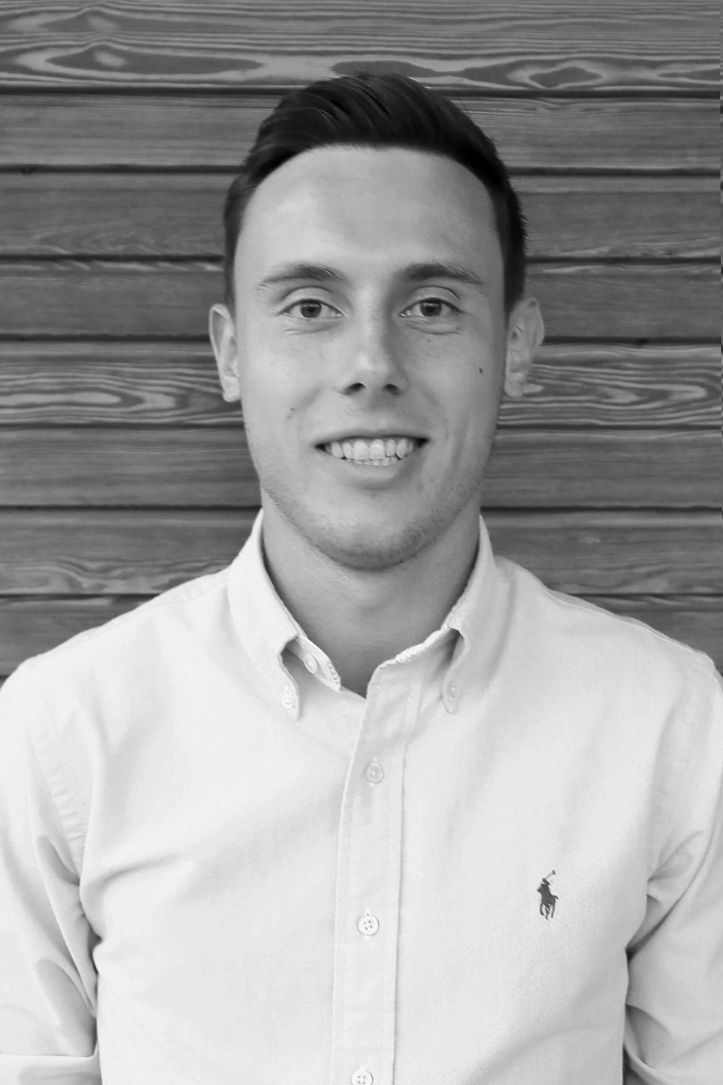 Schwarz-Weiß-Foto eines lächelnden jungen Mannes mit kurzen, gepflegten Haaren, trägt ein helles Button-Down-Hemd mit Polo Ralph Lauren Logo, vor einem Holzpaneel-Hintergrund.