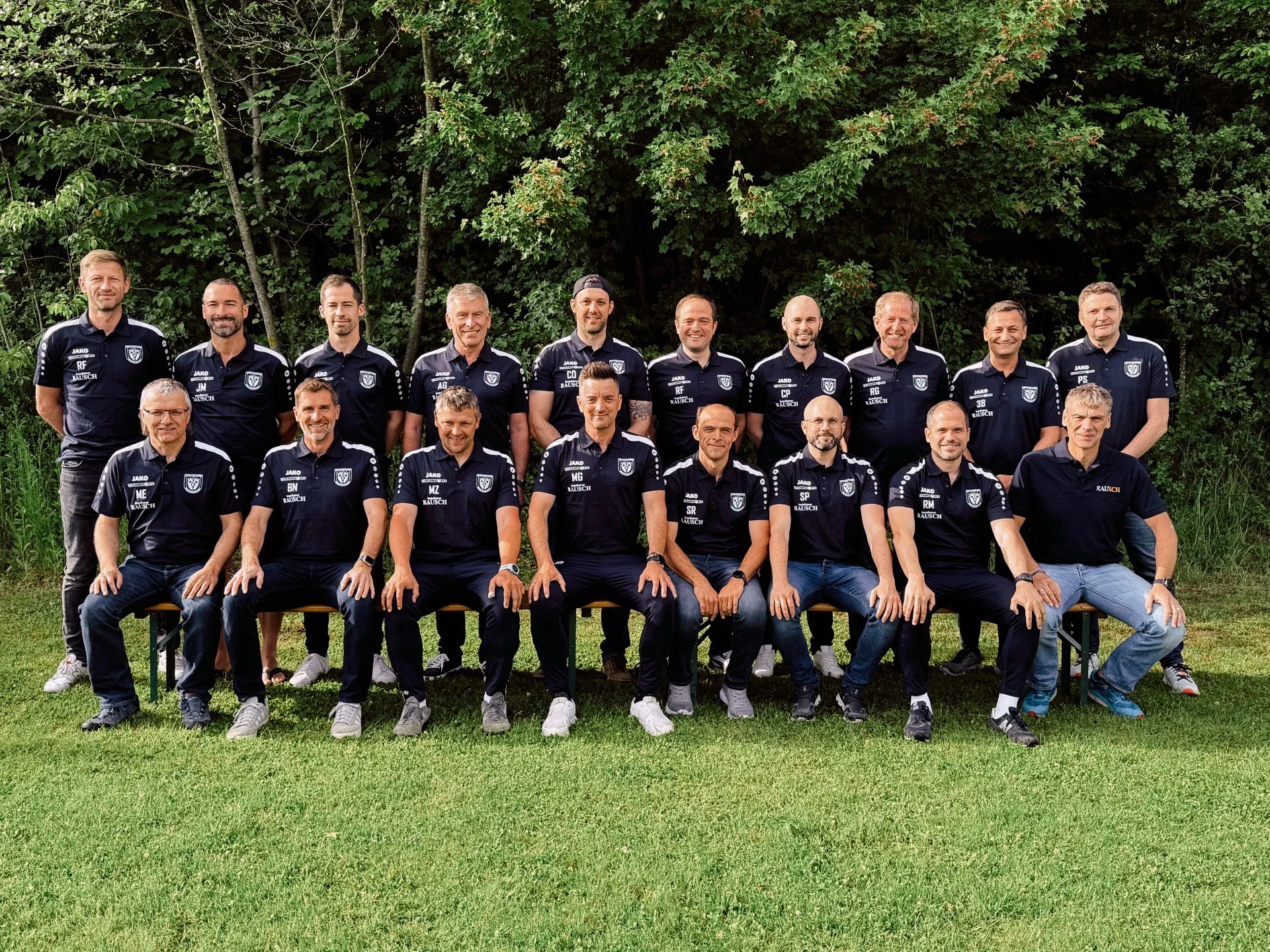 Gruppenum picture von mehreren Männern, die in Fußballtrikots stehen und sitzen, vor einem grünen Hintergrund mit Bäumen.