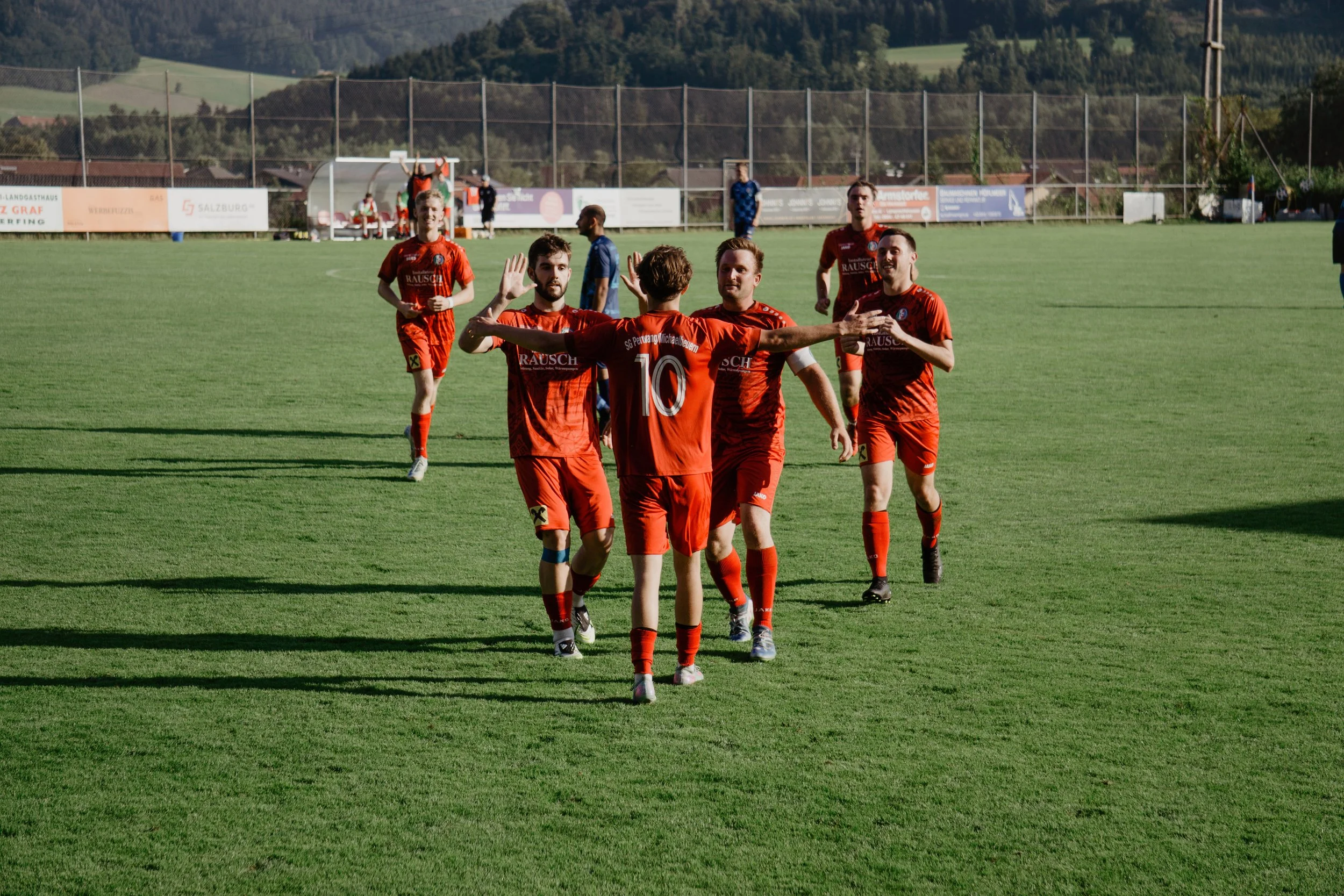 Fußballspieler in roten Trikots feiern auf einem Fußballplatz, umgeben von Hügeln und Bäumen.